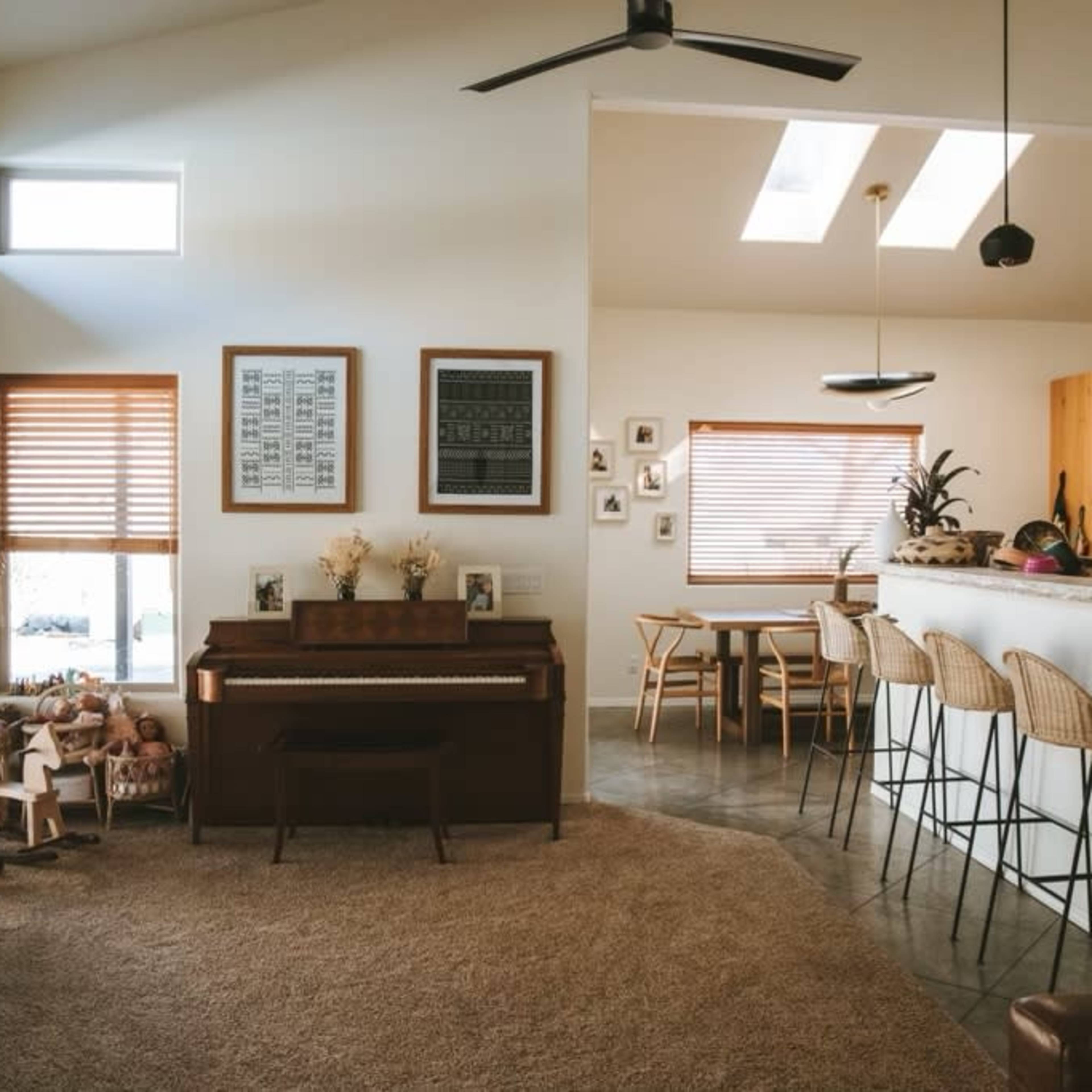 A bright living room with a piano, a dining area with bar stools, and large windows allowing natural light in.