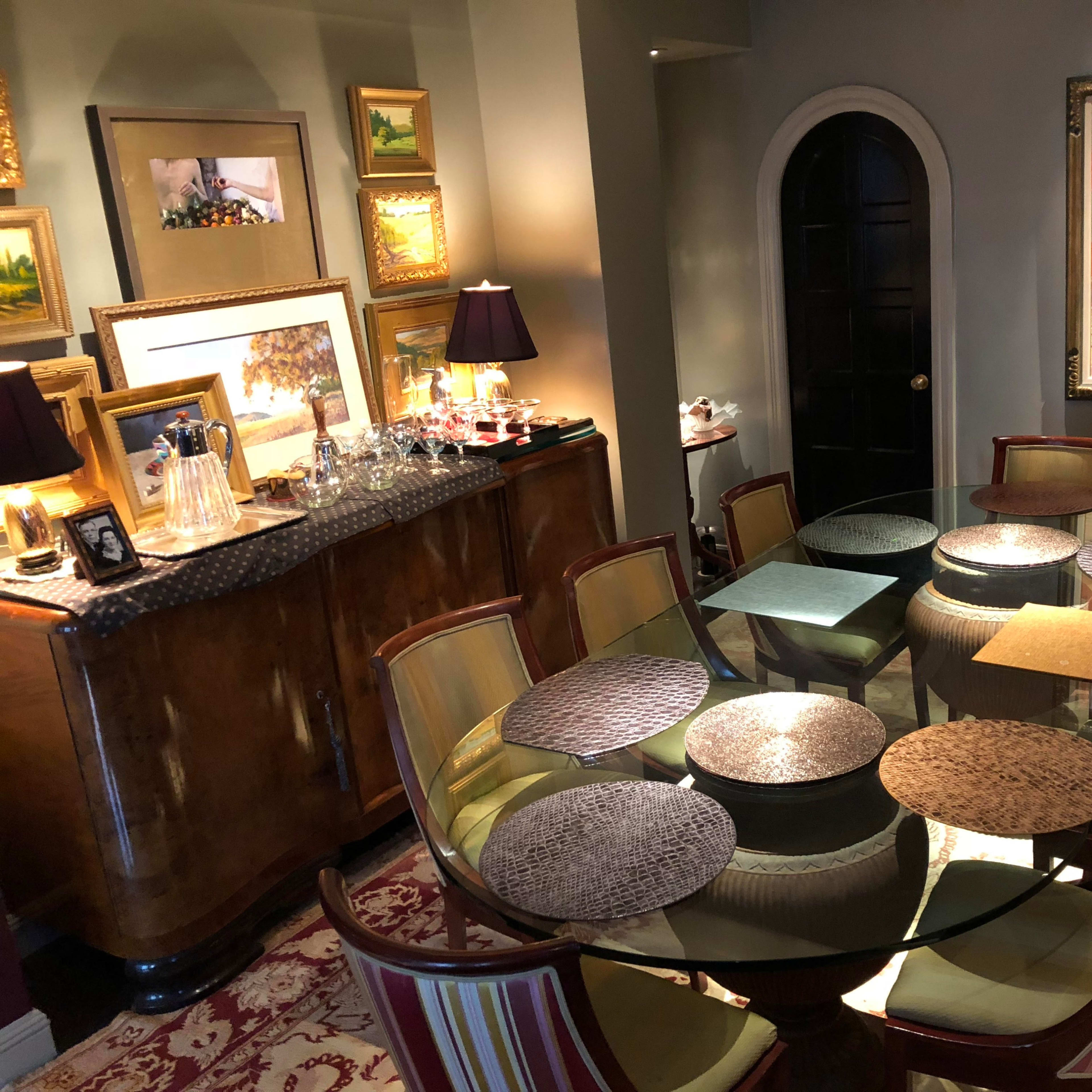 A dining area features a circular glass table surrounded by twelve chairs, with an ornate cabinet holding glassware and artwork adorning the walls.