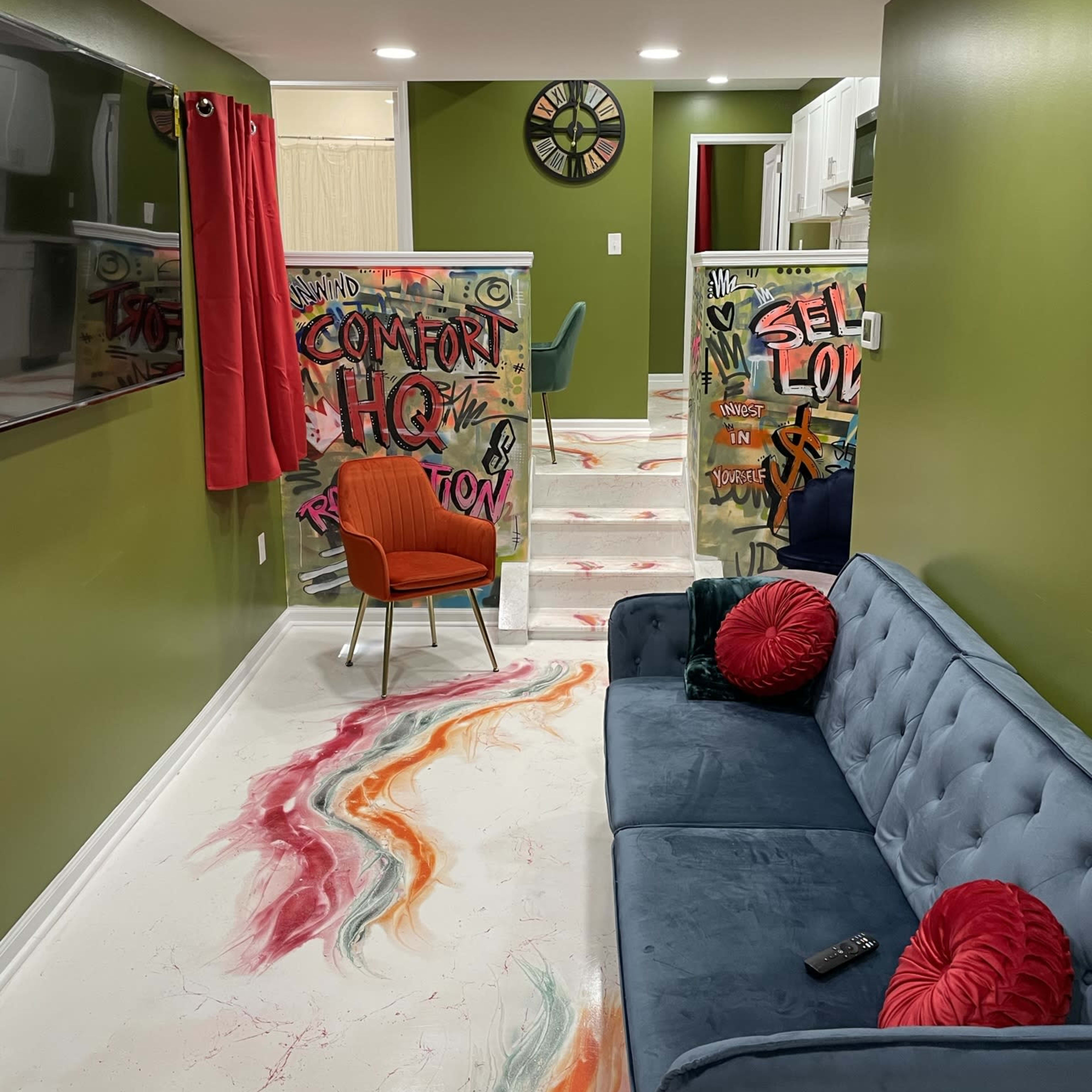 A modern living space with green walls, a blue sofa, colorful graffiti on partitions, and a patterned floor leading to a kitchen area.