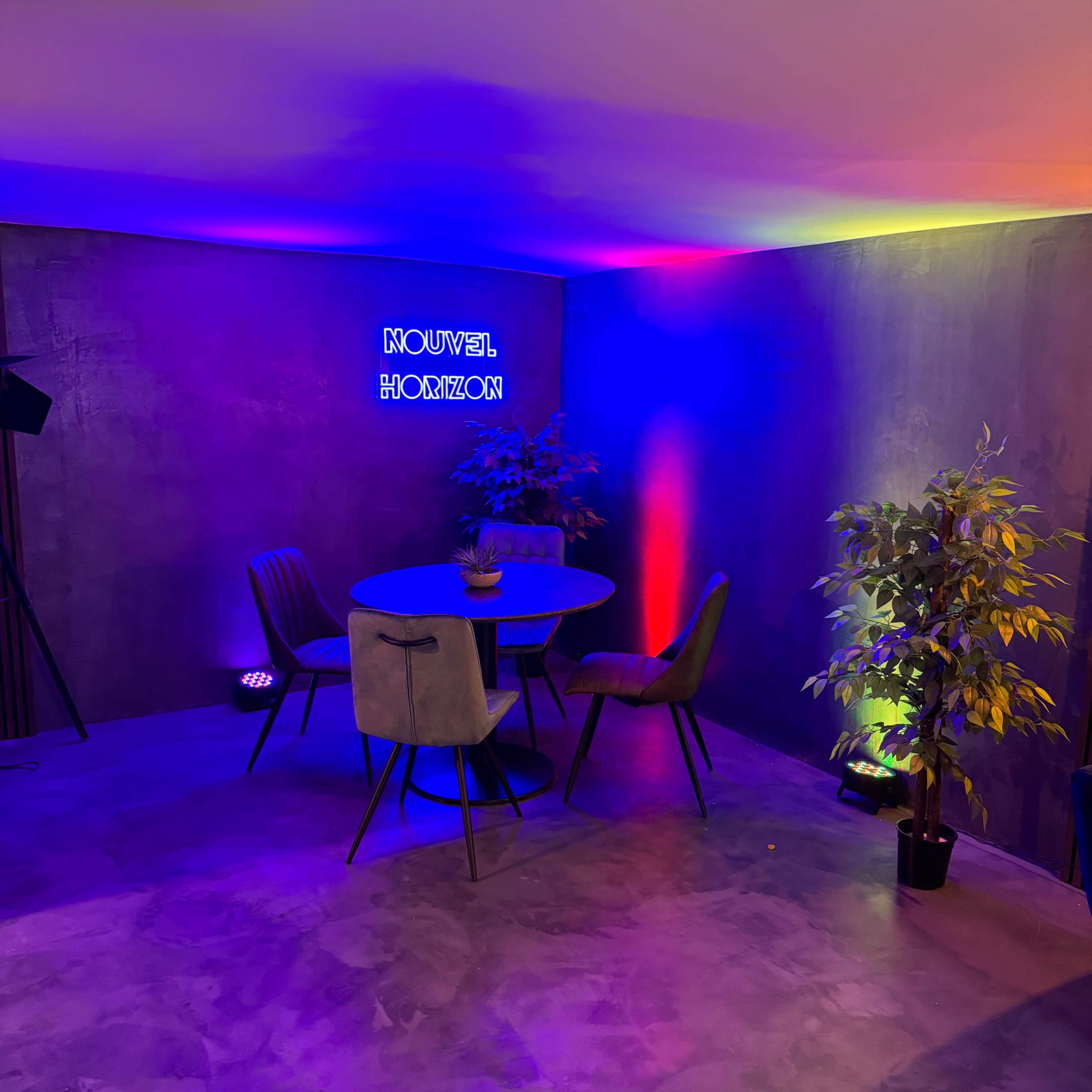 A modern interior space featuring a round table surrounded by four chairs, illuminated by colorful lights and a neon sign.