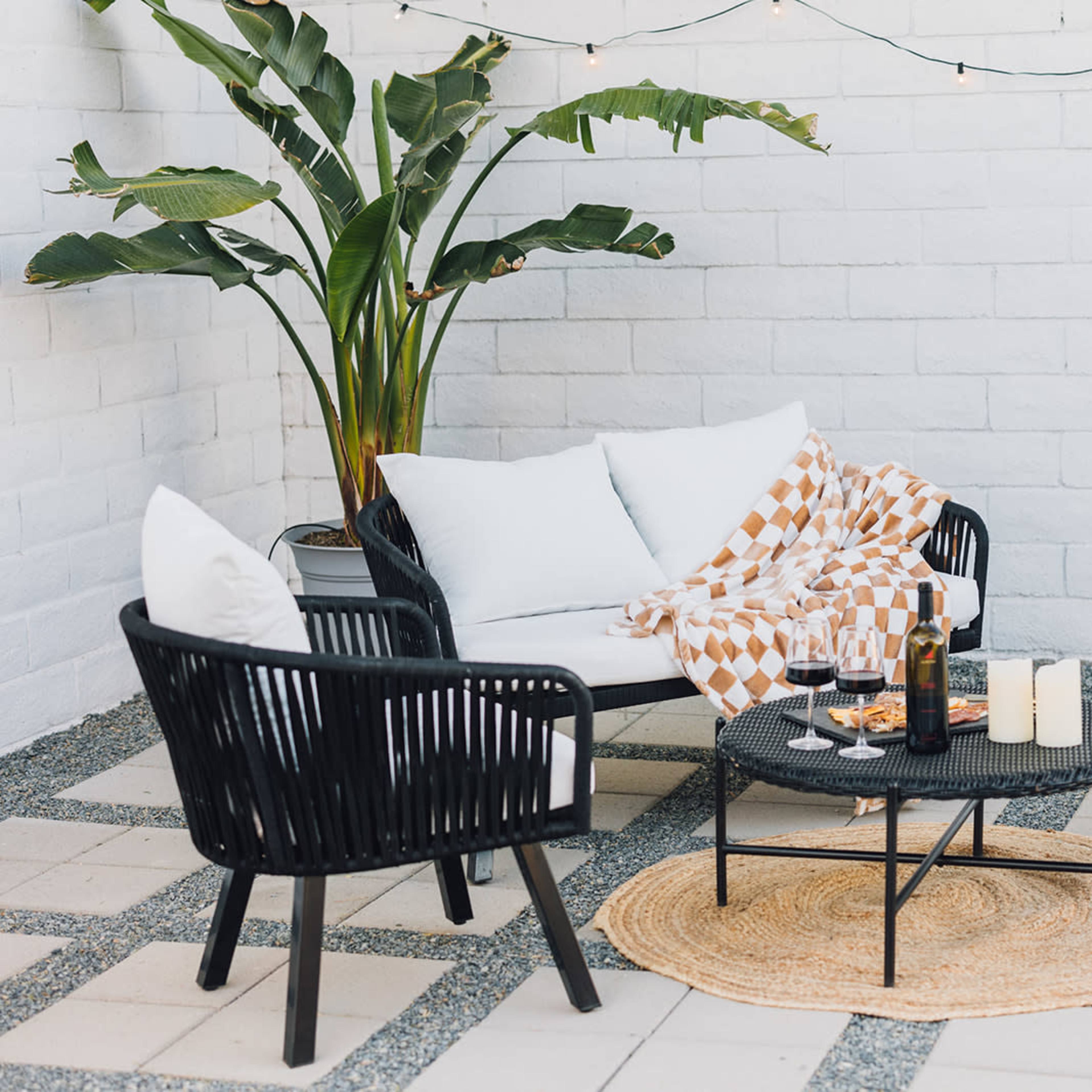 A cozy outdoor seating area with three black wicker chairs, a round table, and a potted plant against a white brick wall, arranged on a patterned patio floor.
