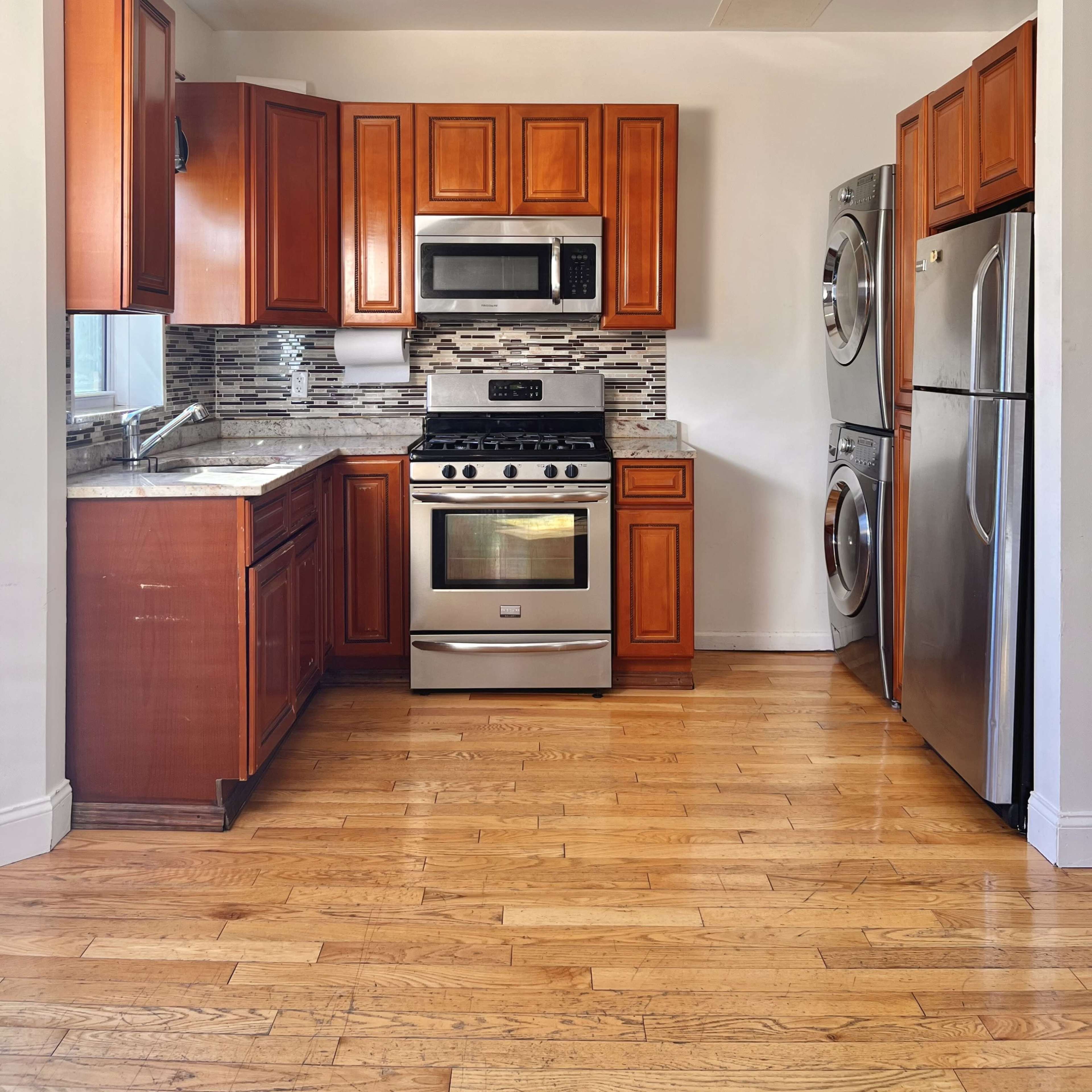 A kitchen with wooden cabinets, a stainless steel stove and microwave, and a washer and dryer stacked next to a refrigerator, all on a hardwood floor.