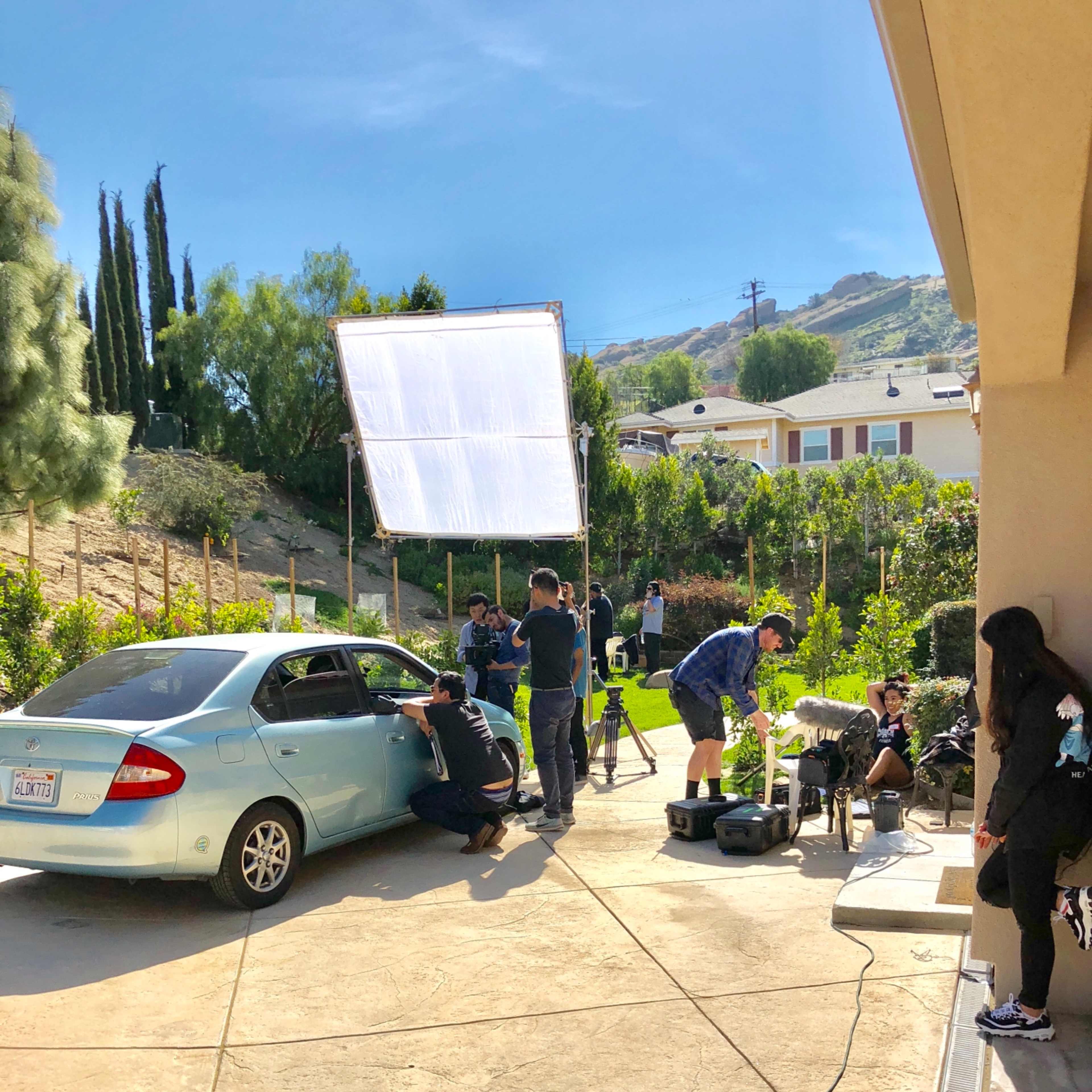 A film crew sets up equipment around a parked car in a garden area with hills in the background.