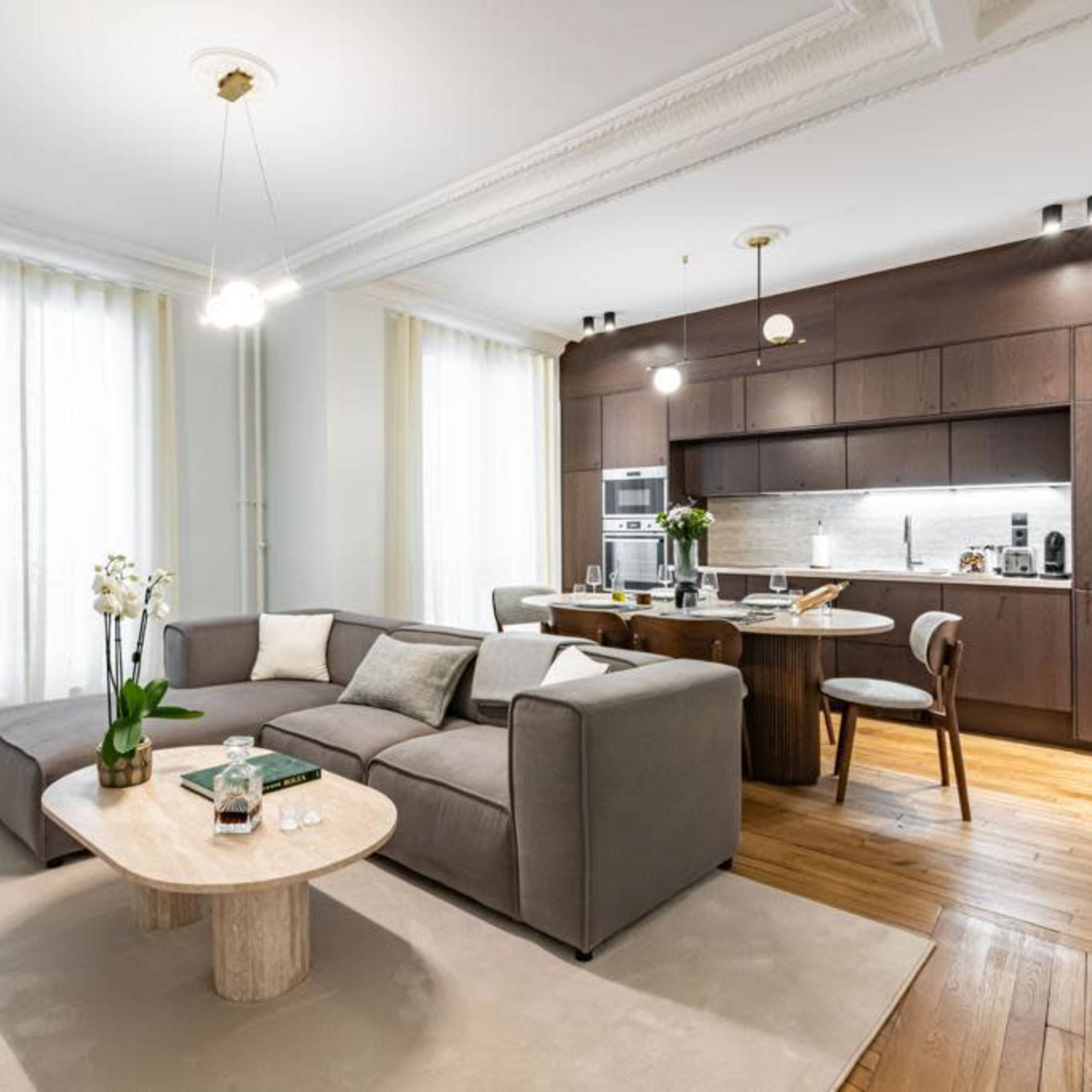 A modern living room features a gray sectional sofa, a round coffee table, and a kitchen area with dark cabinetry and stainless steel appliances.
