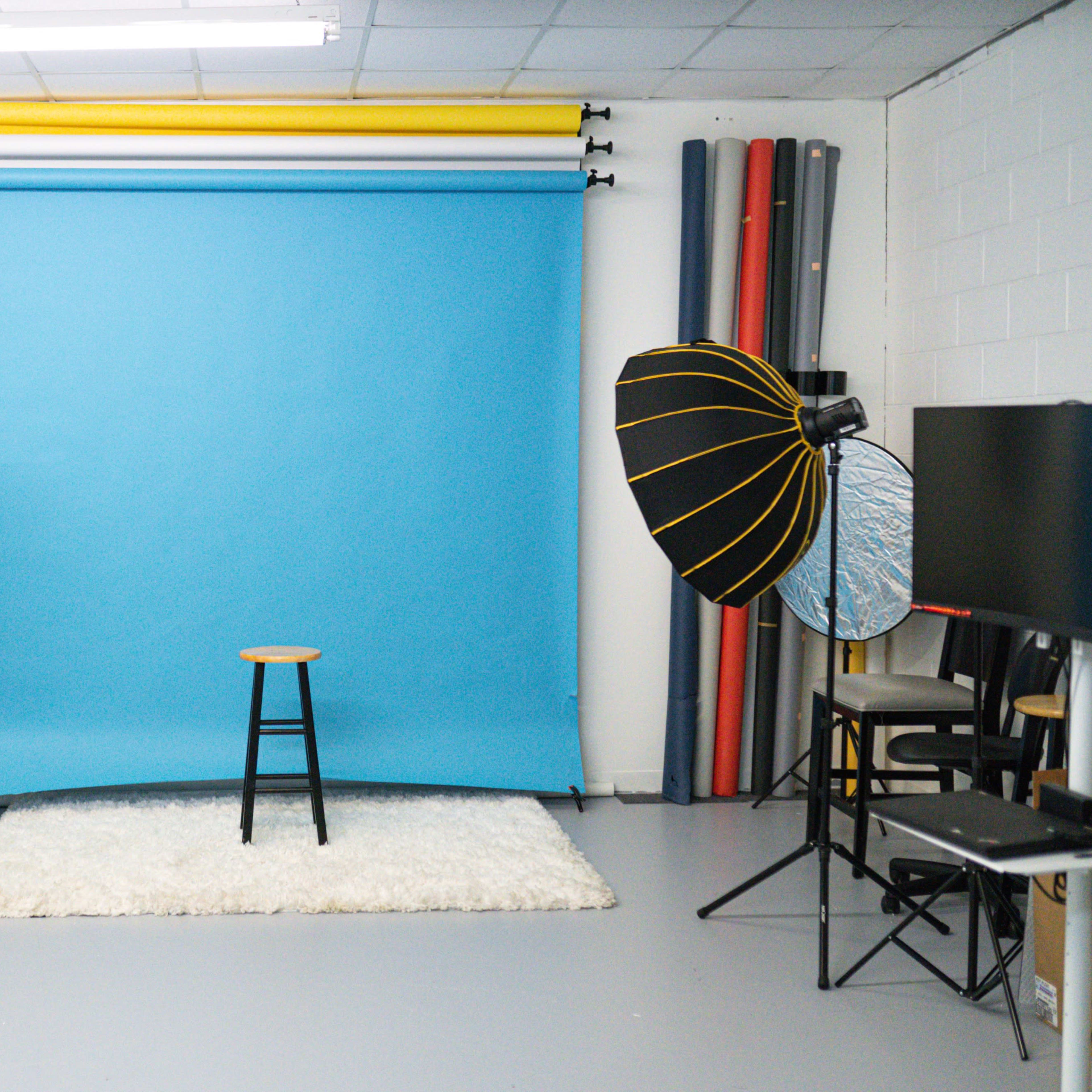 A photography studio equipped with a blue backdrop, two softbox lights, a stool, and a rug.
