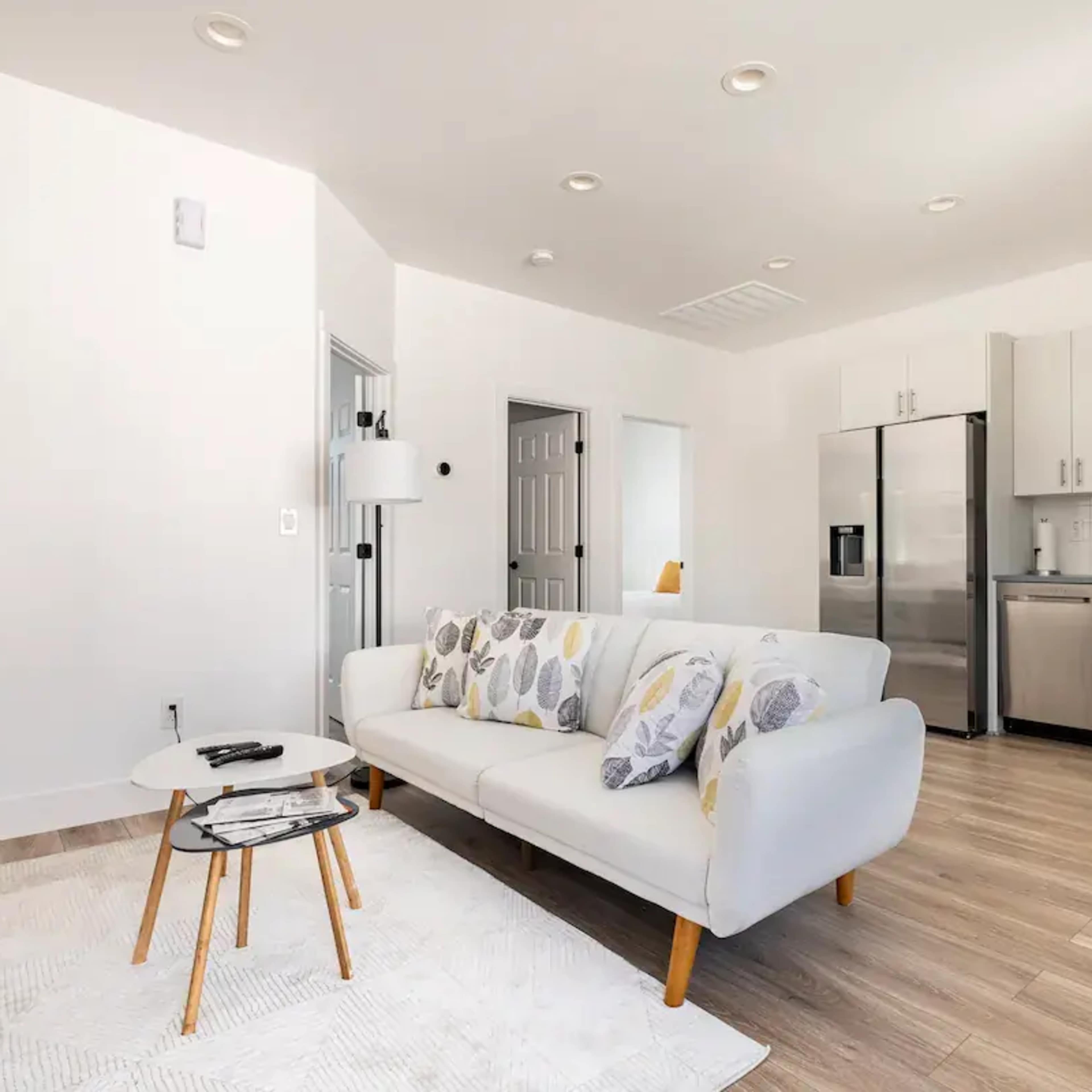 A modern living room features a white sofa with decorative cushions, a small coffee table, and an open kitchen area with stainless steel appliances.