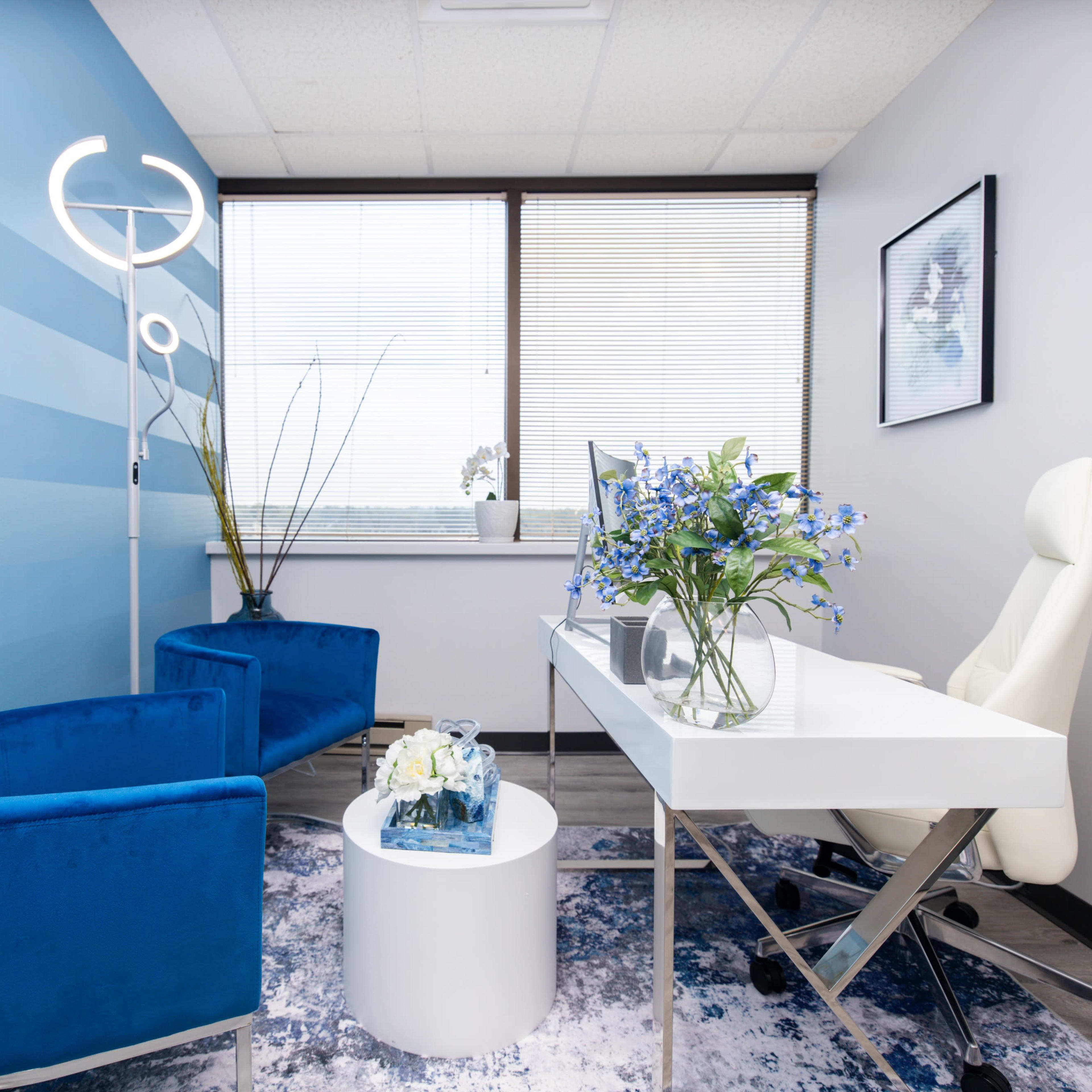 A modern office space with blue accent walls, two blue chairs, a white desk, and a vase of flowers on a round table.