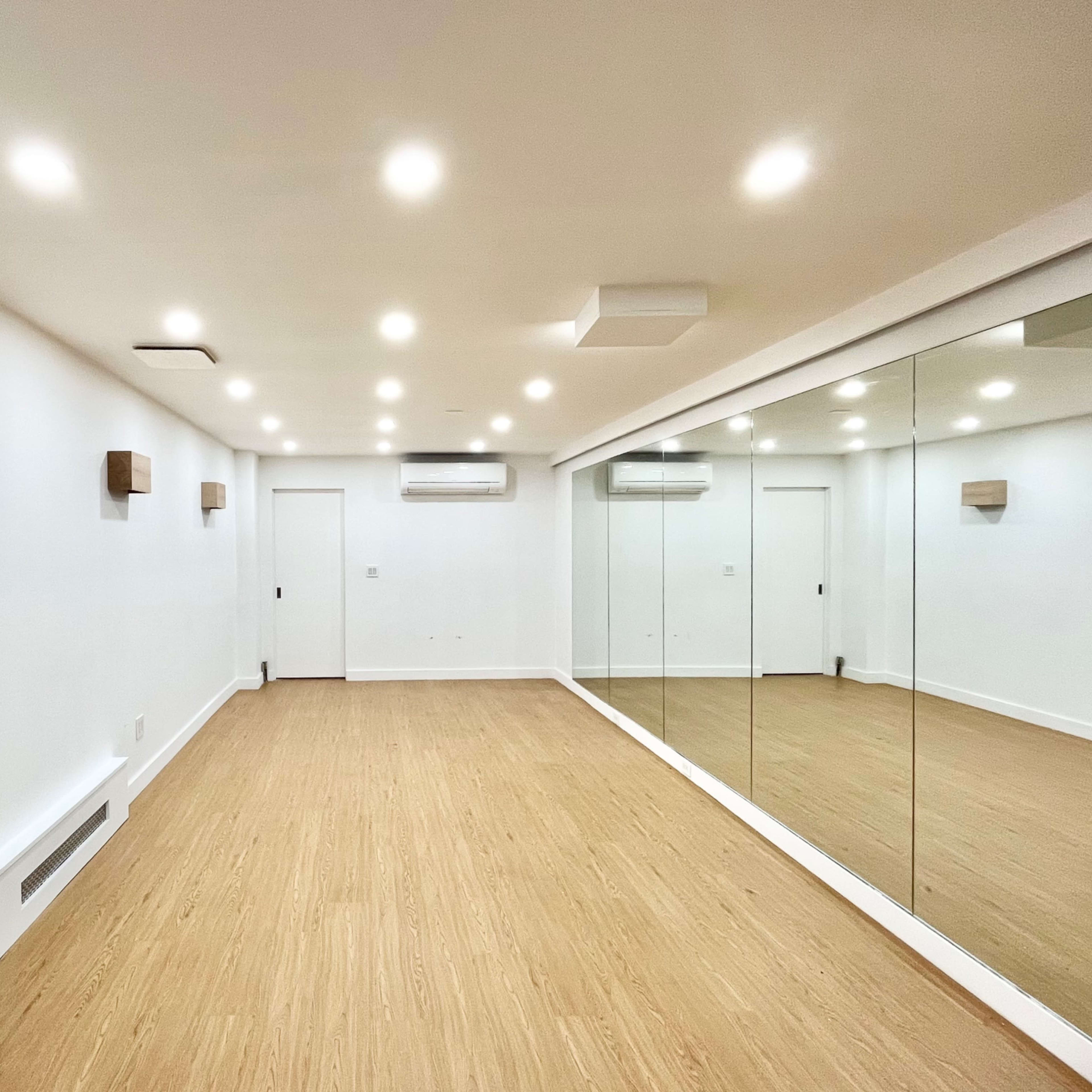 A minimalist room with light wood flooring and a mirrored wall, featuring recessed lighting and minimal wall decor.