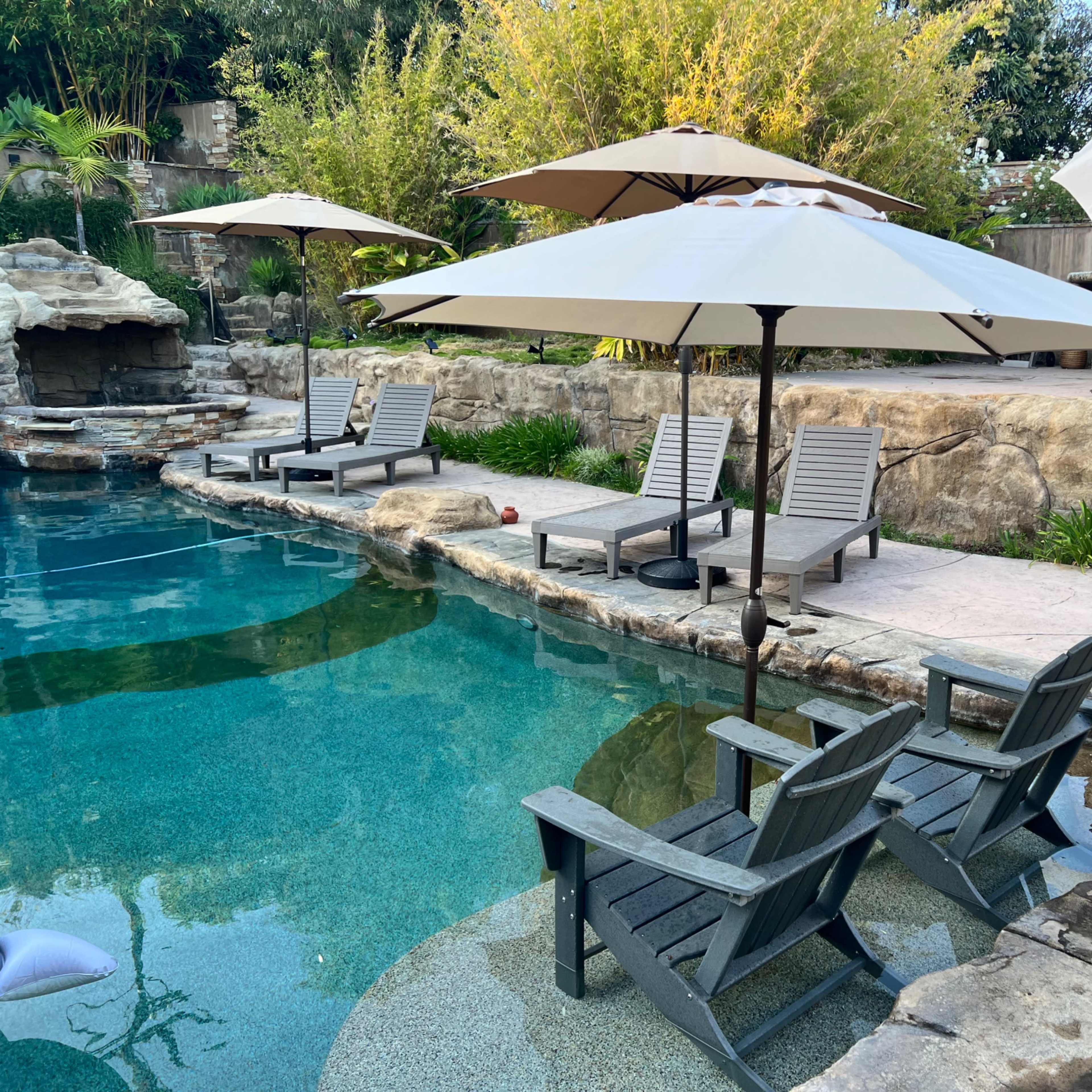 A backyard pool area with lounge chairs and umbrellas surrounding a clear water pool, complemented by rocky landscaping and green plants.