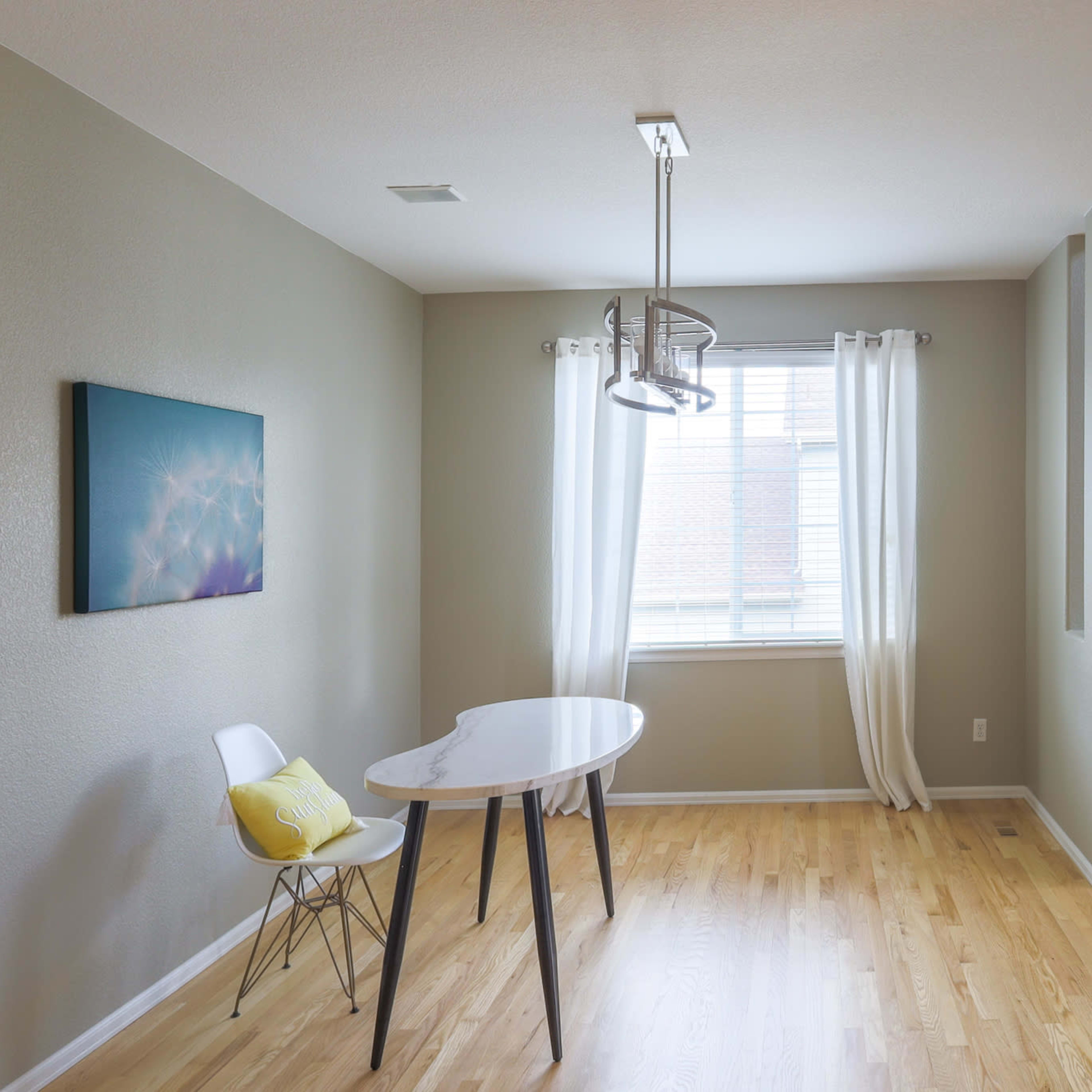 A minimalistic interior featuring a small table with a white chair, a light fixture, a window with sheer curtains, and a modern armchair in a corner.