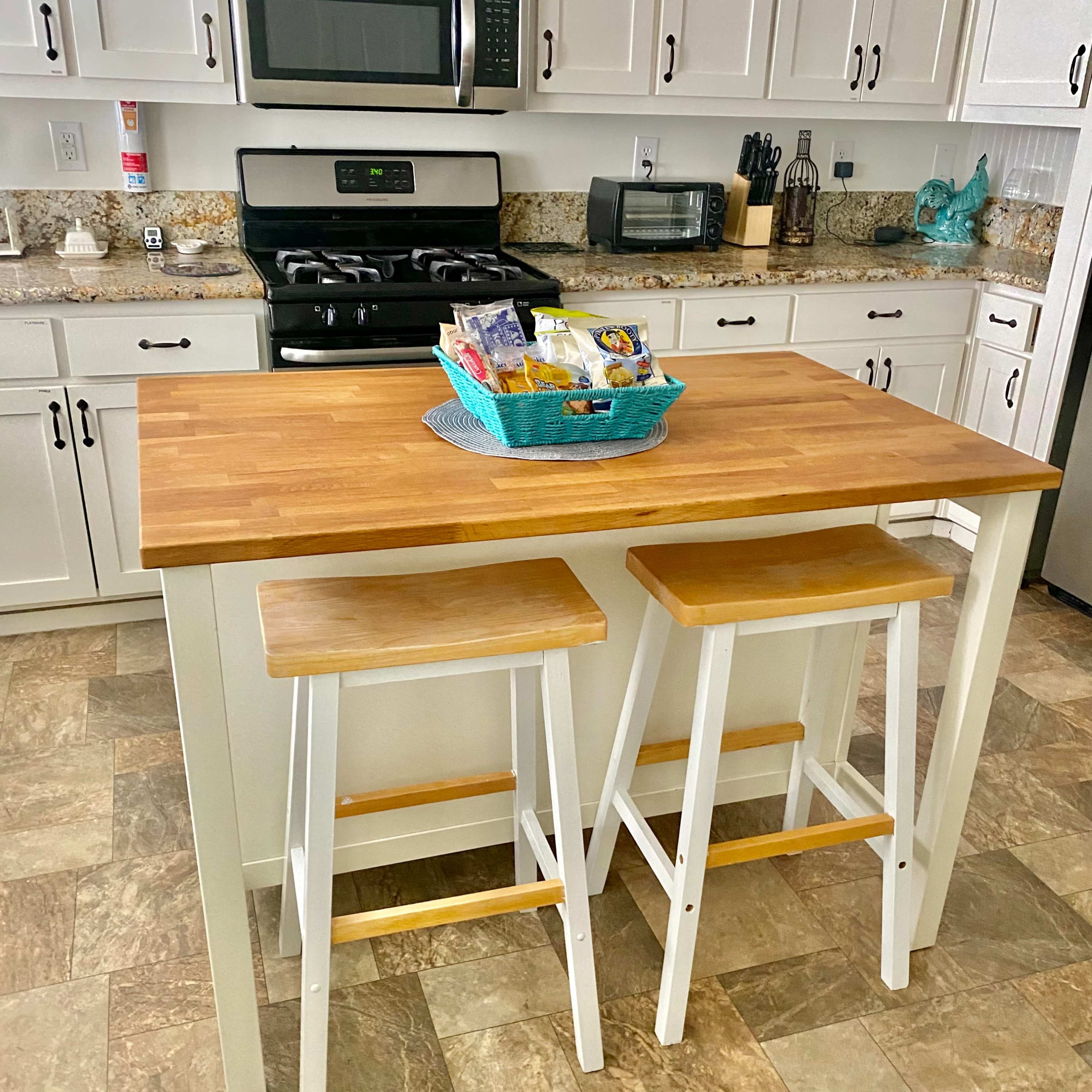 A wooden kitchen island with two matching stools sits in a modern kitchen featuring granite countertops and stainless steel appliances.