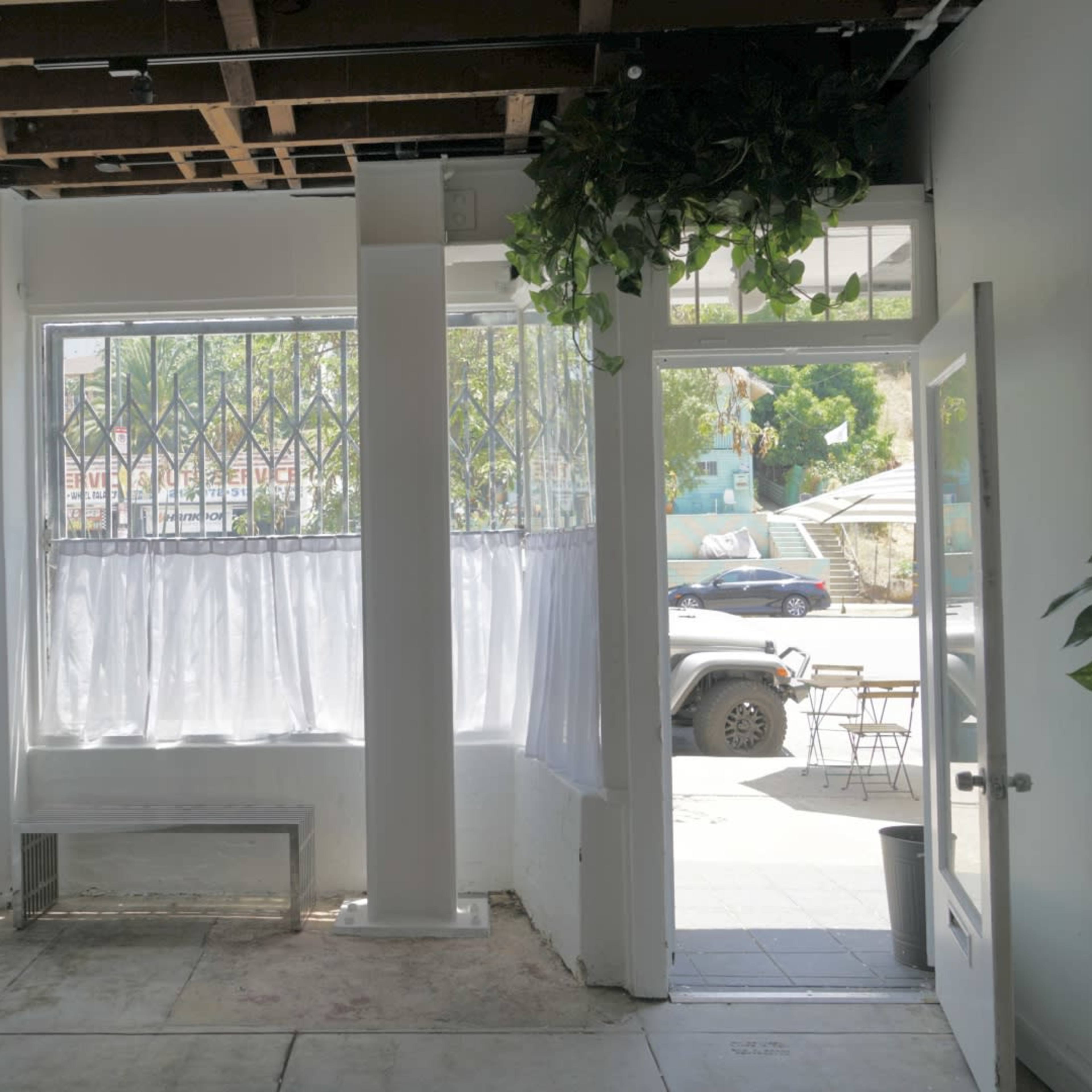 A small, empty interior space with white walls, a large window covered by sheer curtains, and an open door leading outside.