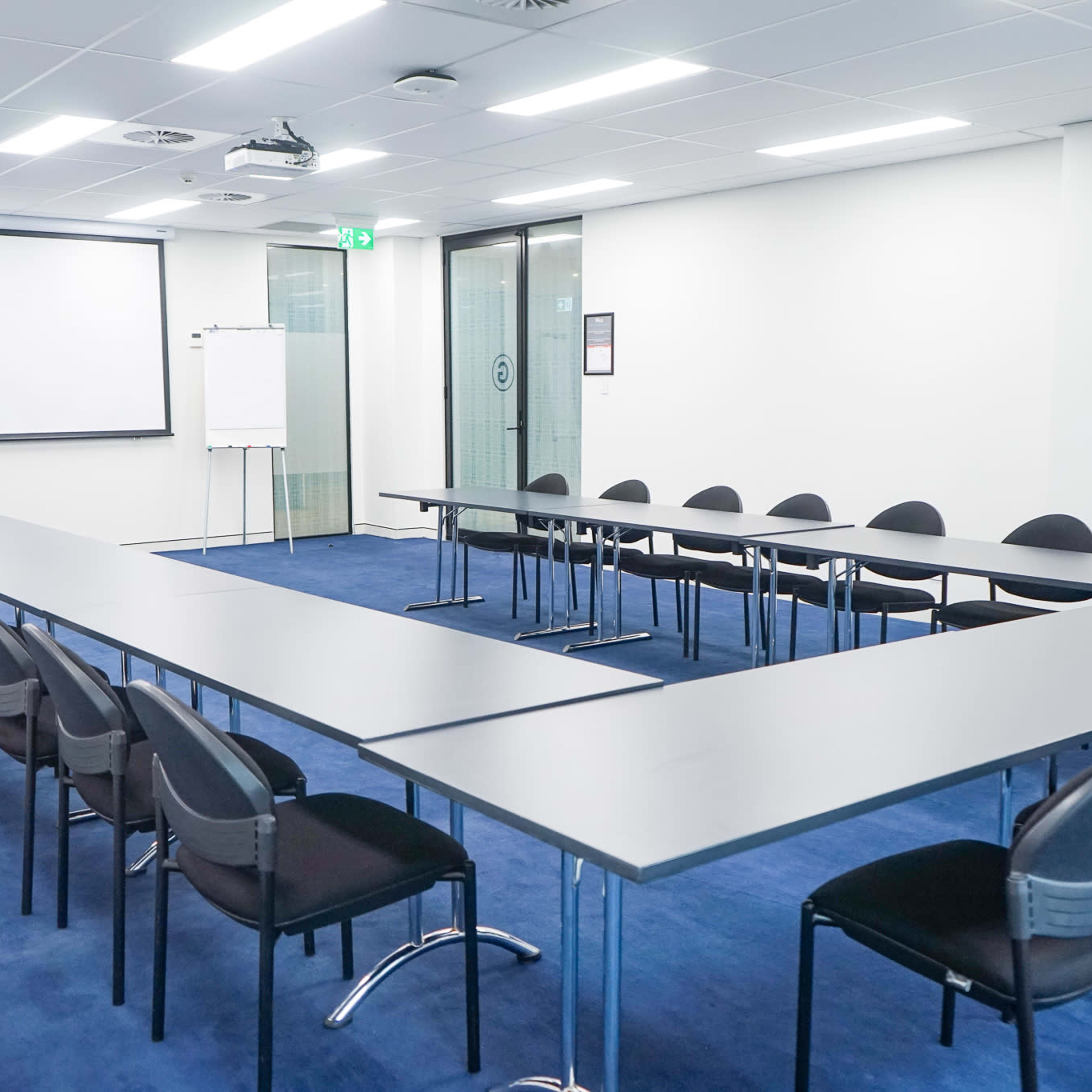 A spacious conference room features a long arrangement of tables and black chairs, with a projector screen and whiteboards at one end.