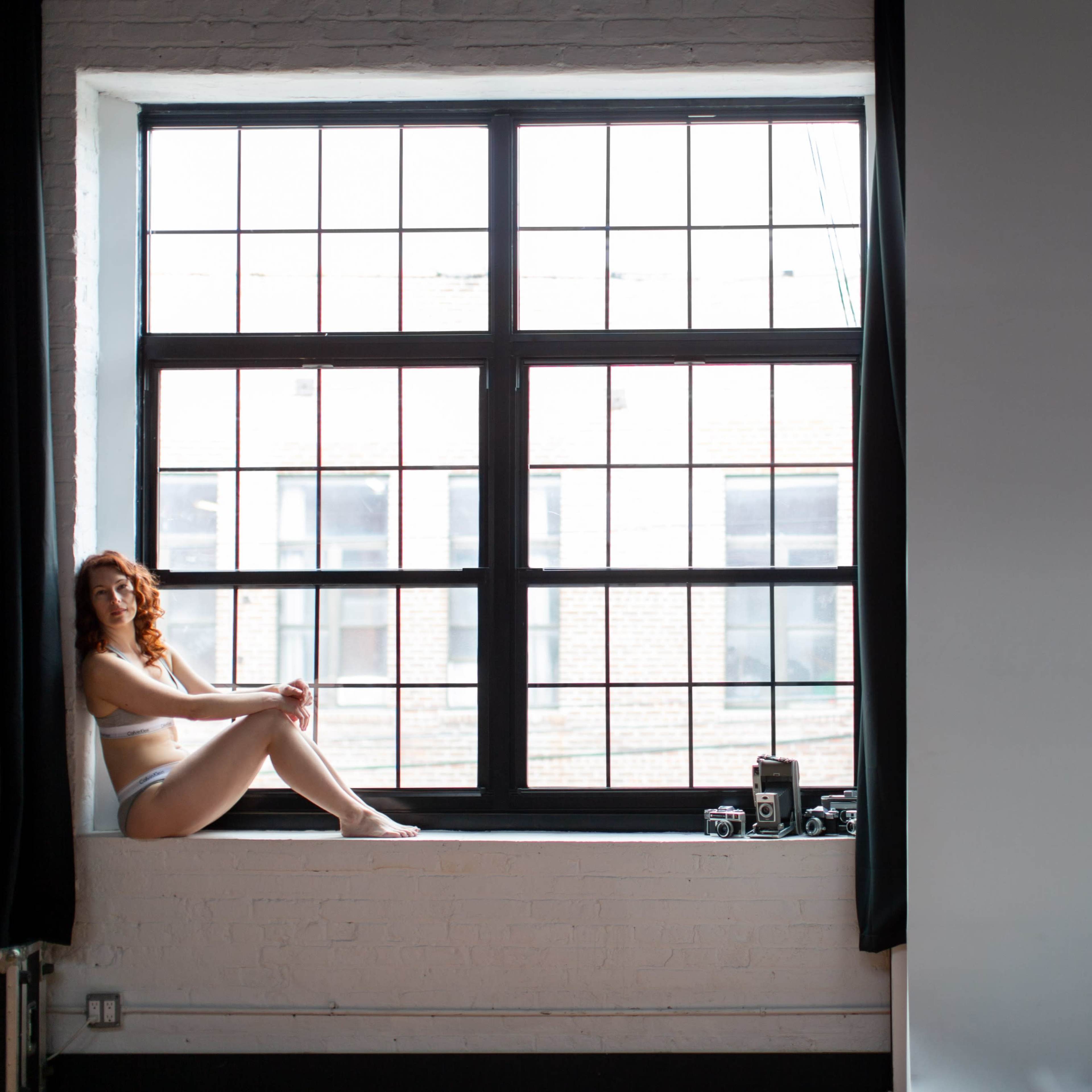 A person is sitting on a windowsill in a bright room with large, multi-paned windows.
