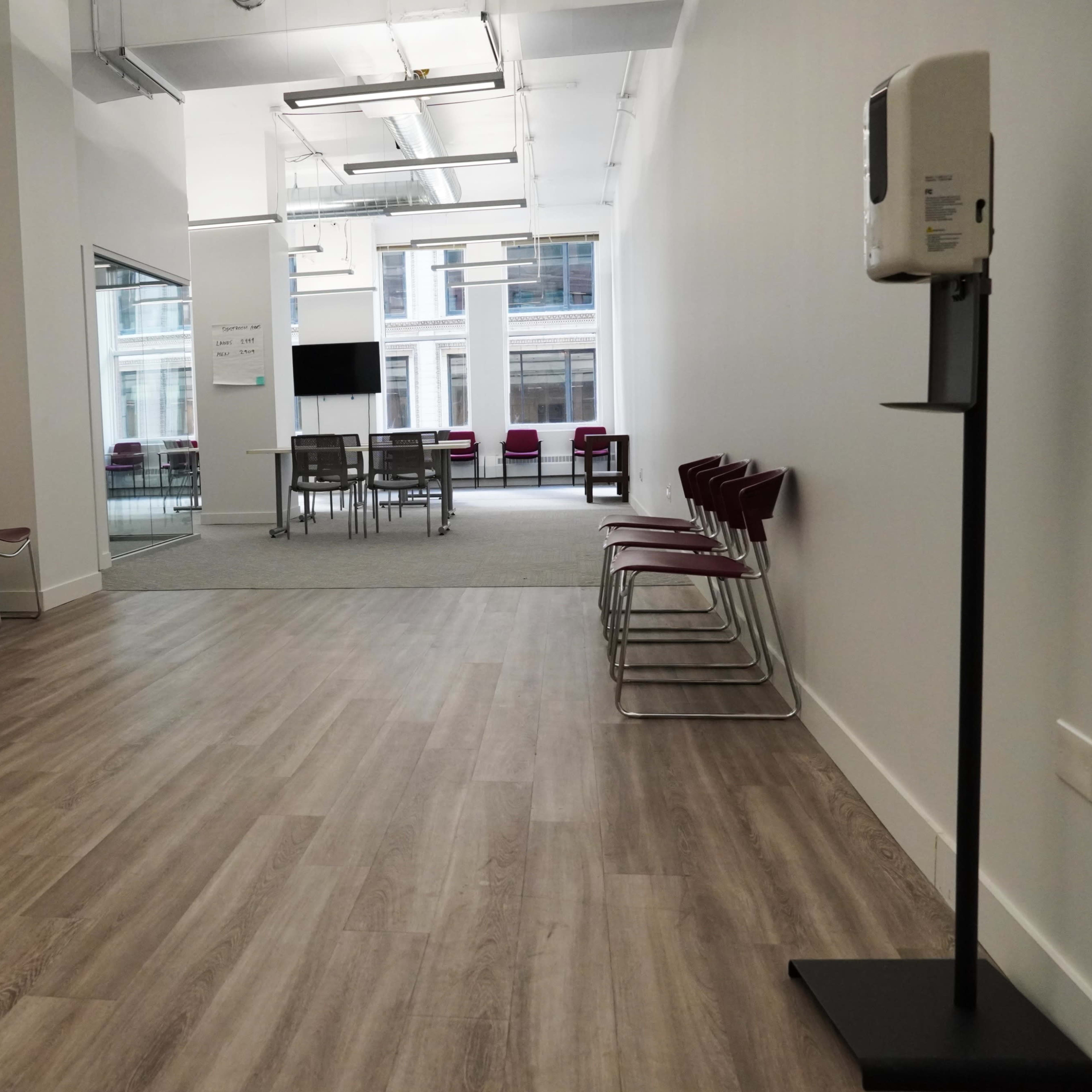 A minimalist waiting area features a row of metal chairs on one side and a glass-walled meeting space in the background.