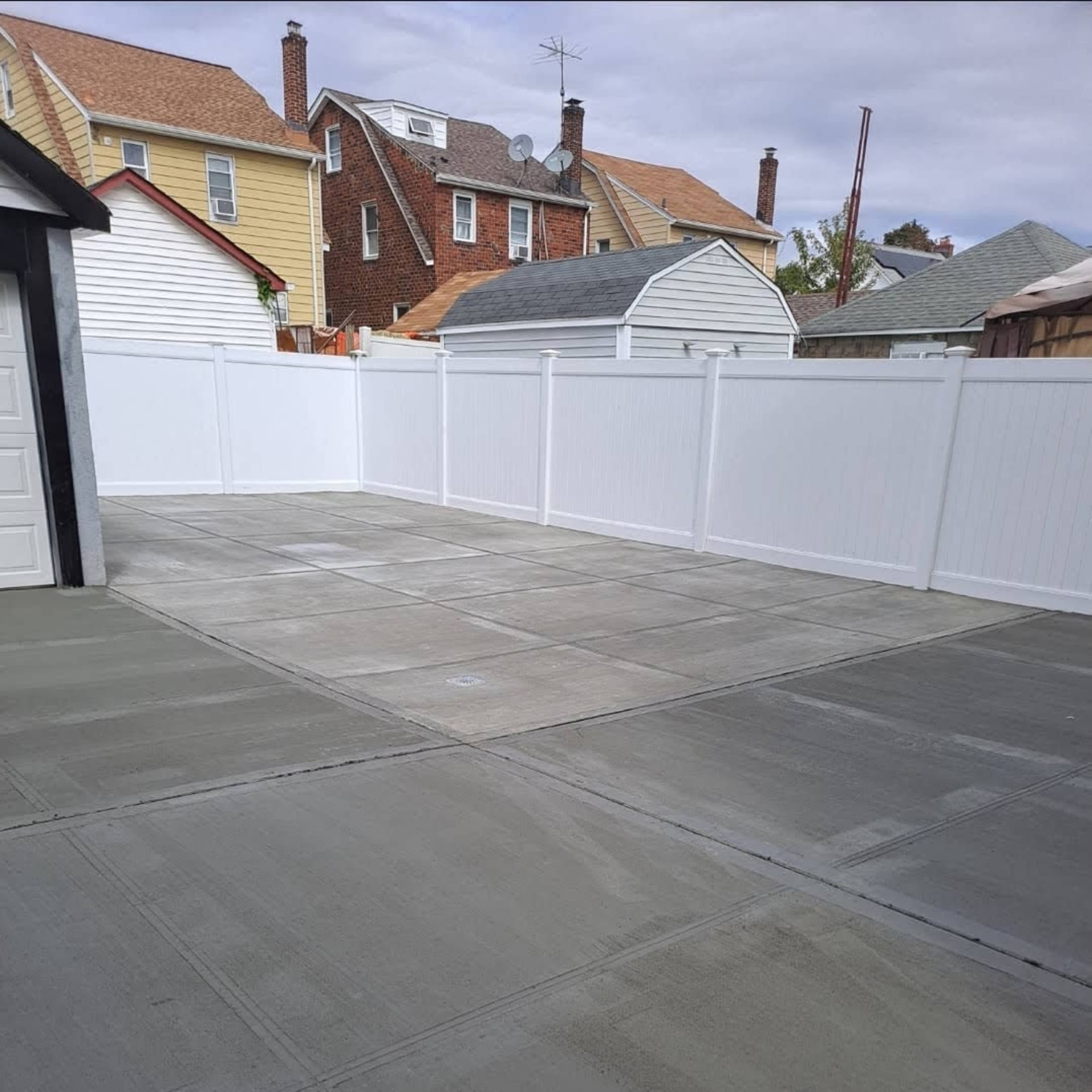 A newly poured concrete surface in a backyard, enclosed by a white fence, with several houses visible in the background.