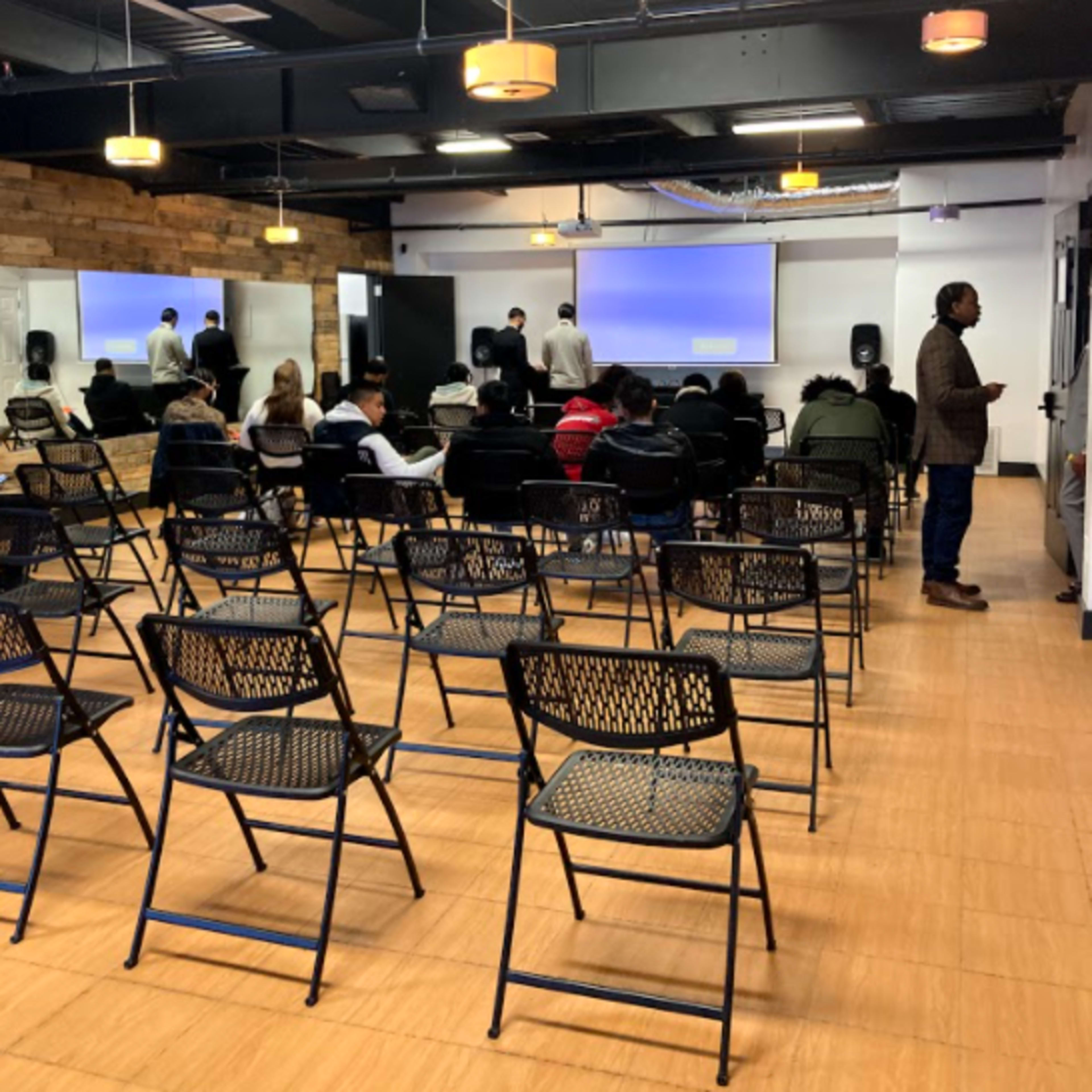 A crowded seminar room with rows of empty chairs facing a presentation screen, while a few individuals stand near the front.