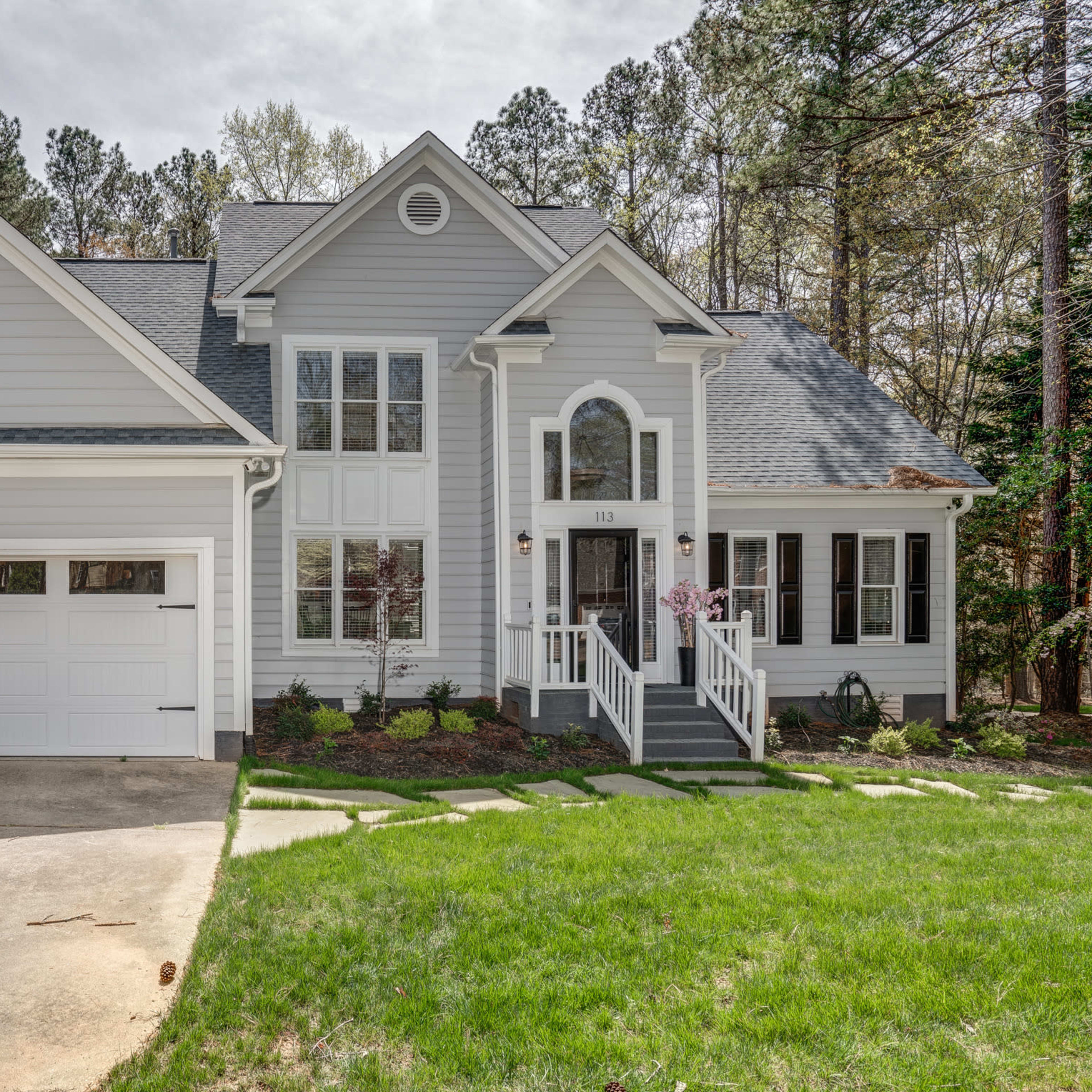 A single-story house with a gray exterior, symmetrical windows, and a two-car garage is set against a backdrop of trees and greenery.