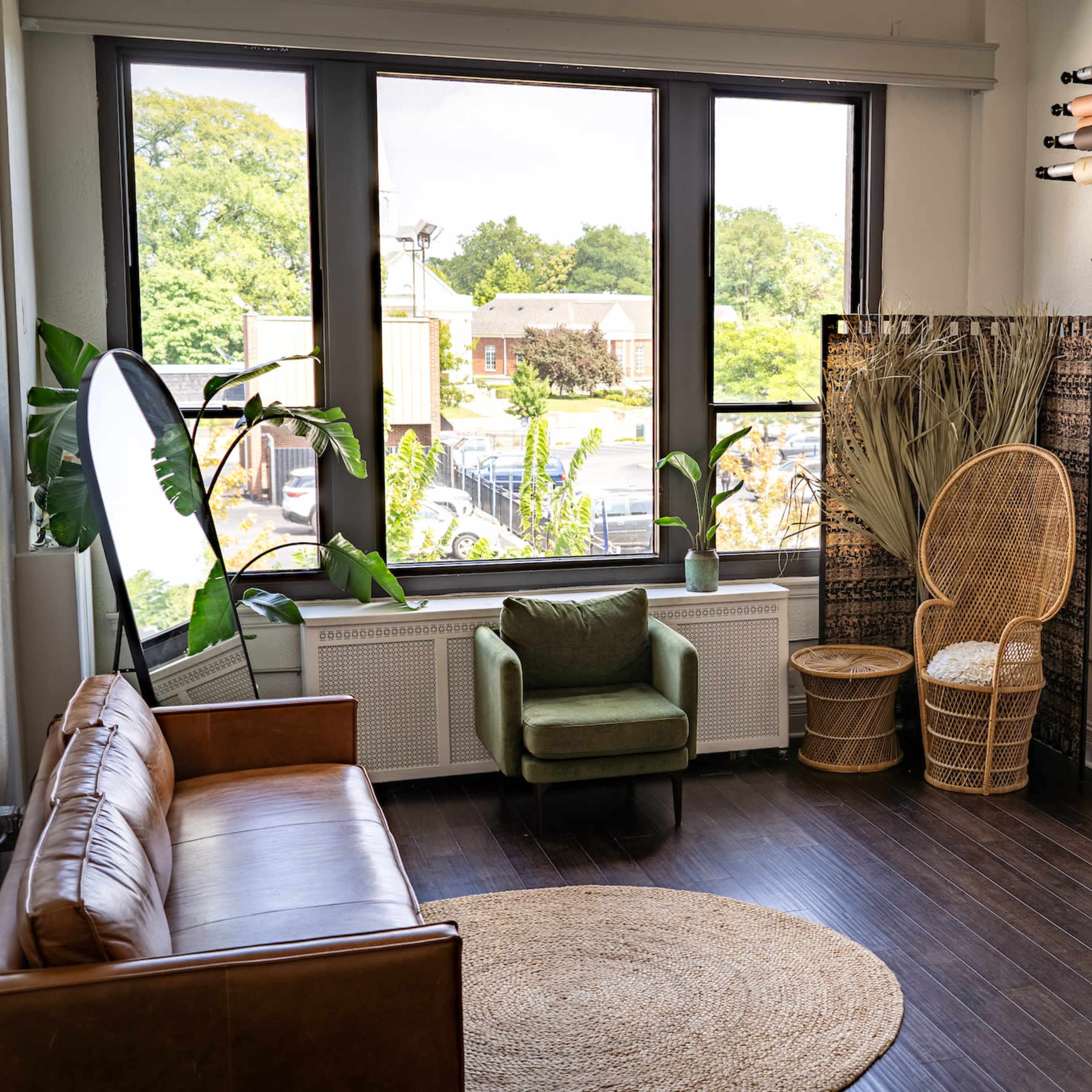 A modern studio setup featuring a leather couch, a green armchair, a round rug, a wicker chair, and a large window with natural light.