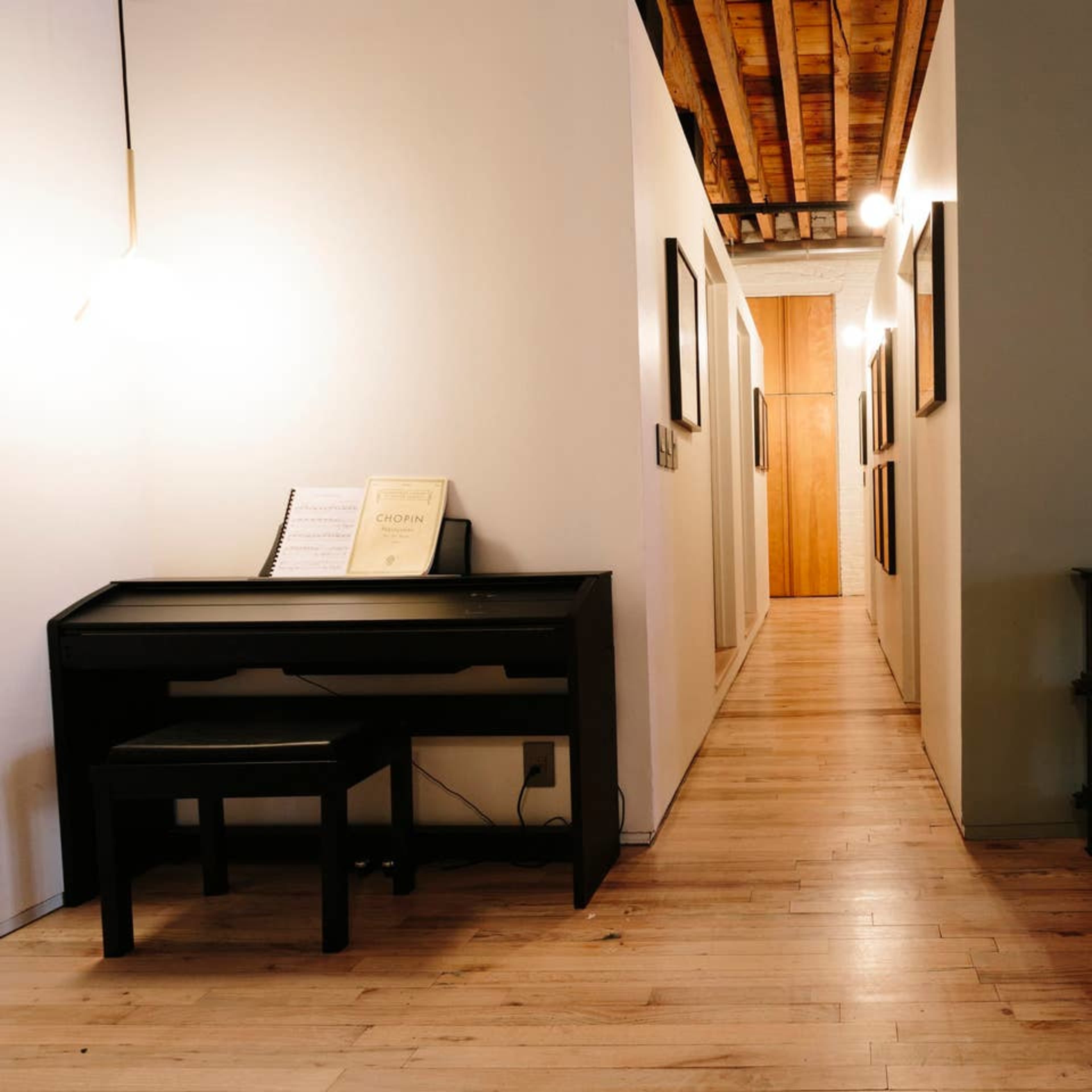 A long, narrow hallway with wooden floors, featuring a black desk and stool on the left and framed artworks along the walls, leading to a light-colored doorway.