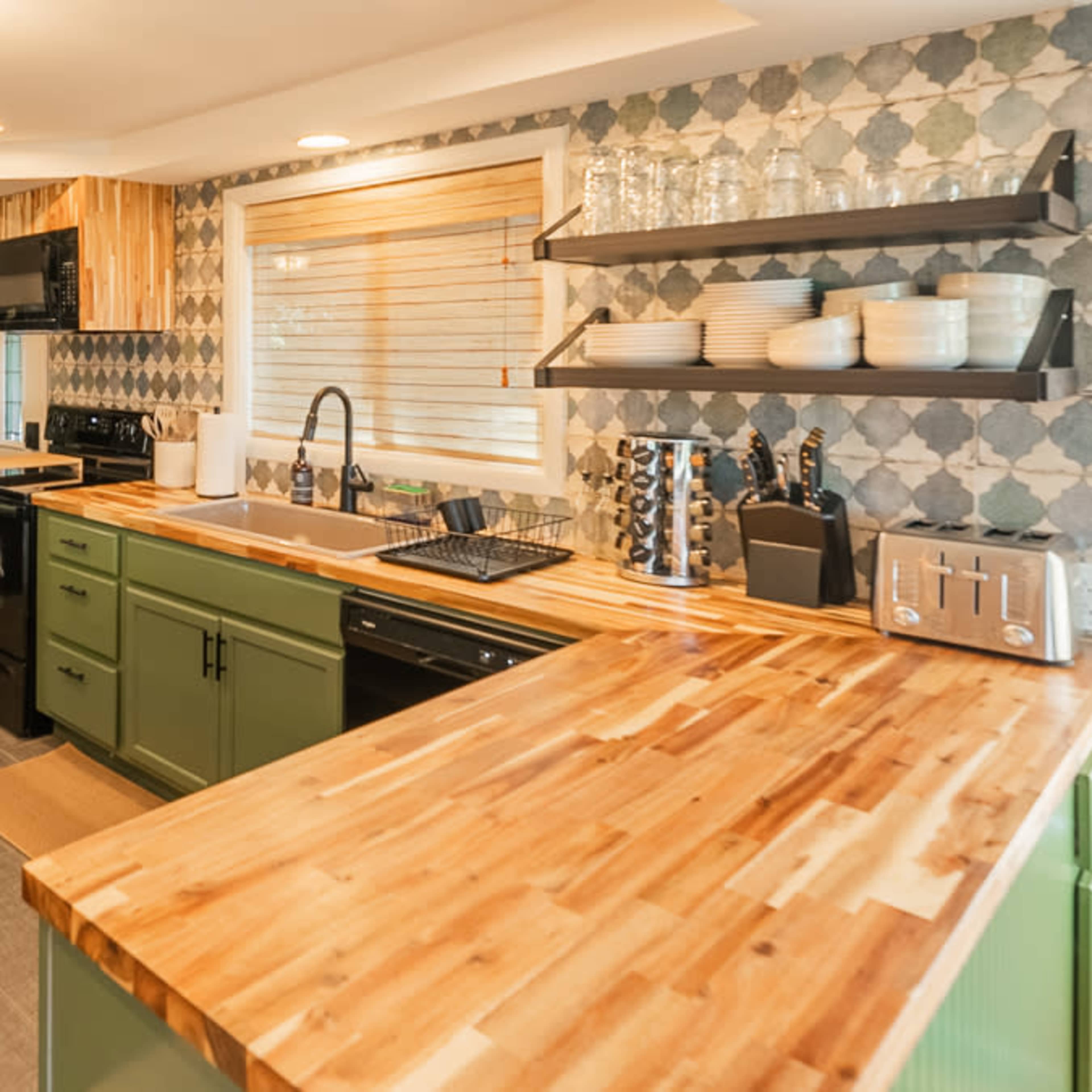 A modern kitchen features green cabinets, a wooden countertop, and open shelving with dishware against a patterned backsplash.