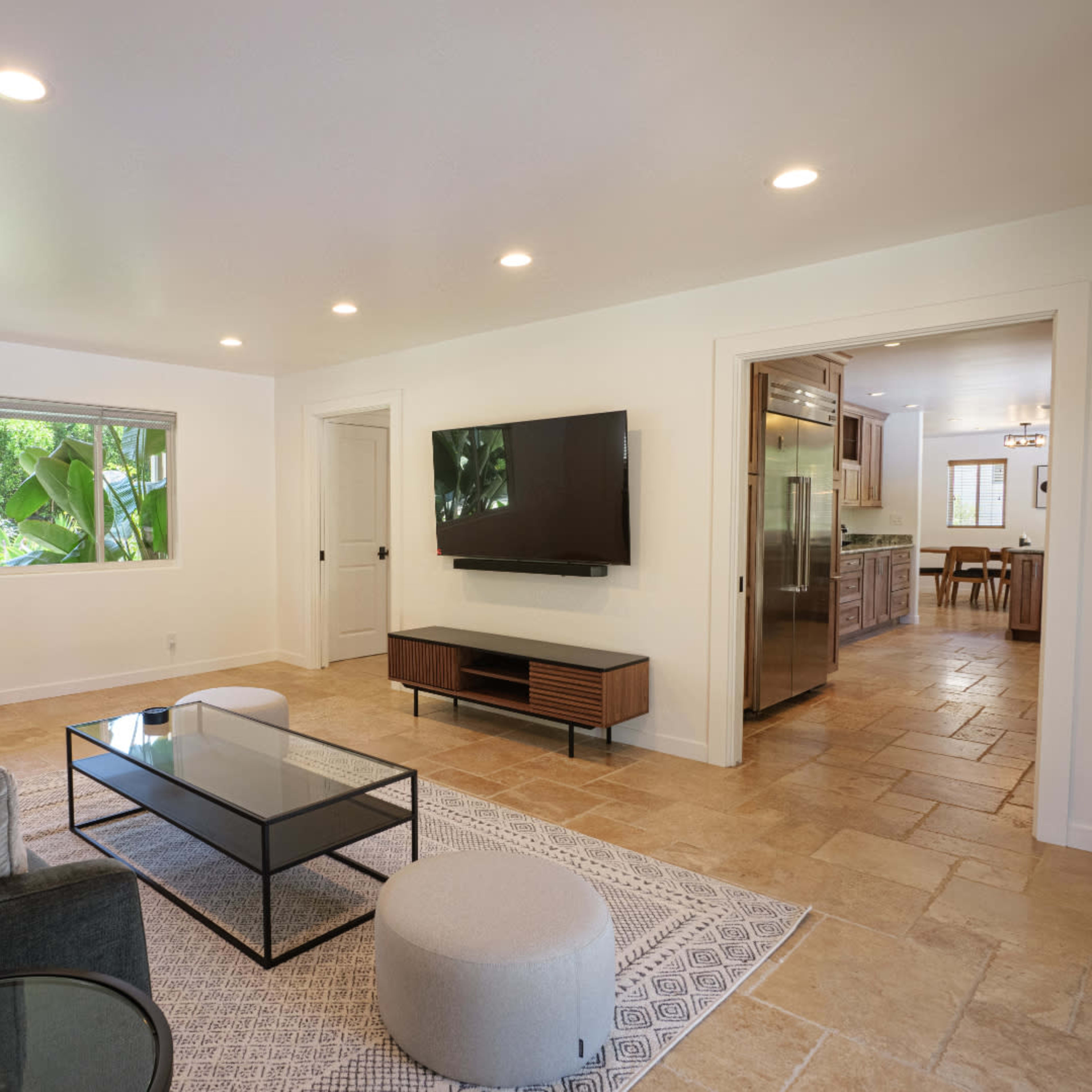 A modern living room features a gray sofa, a glass coffee table, a round pouf, and a wall-mounted television, with a window showcasing greenery outside.