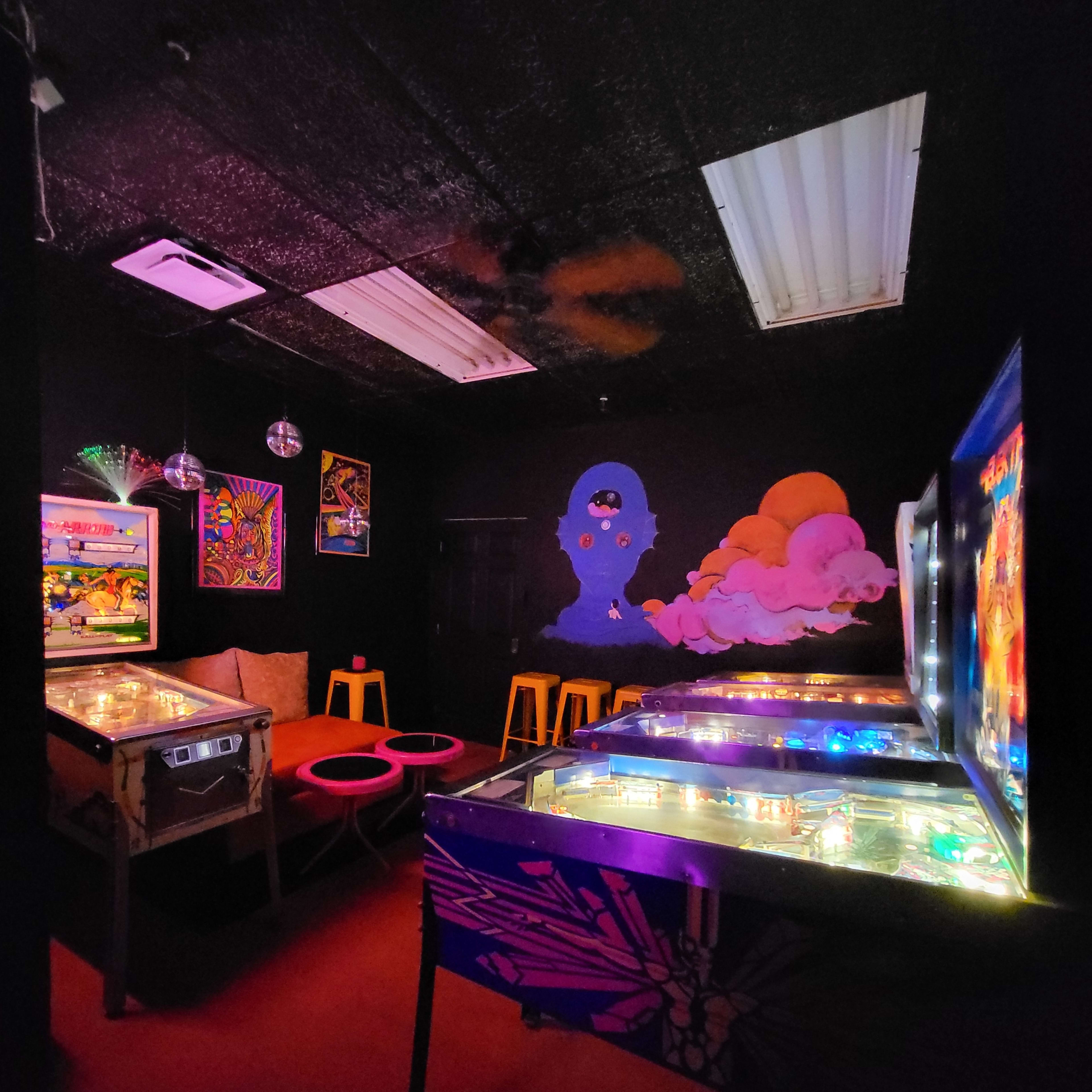 A dimly lit arcade room with colorful pinball machines, vibrant wall art, and a seating area with red stools.