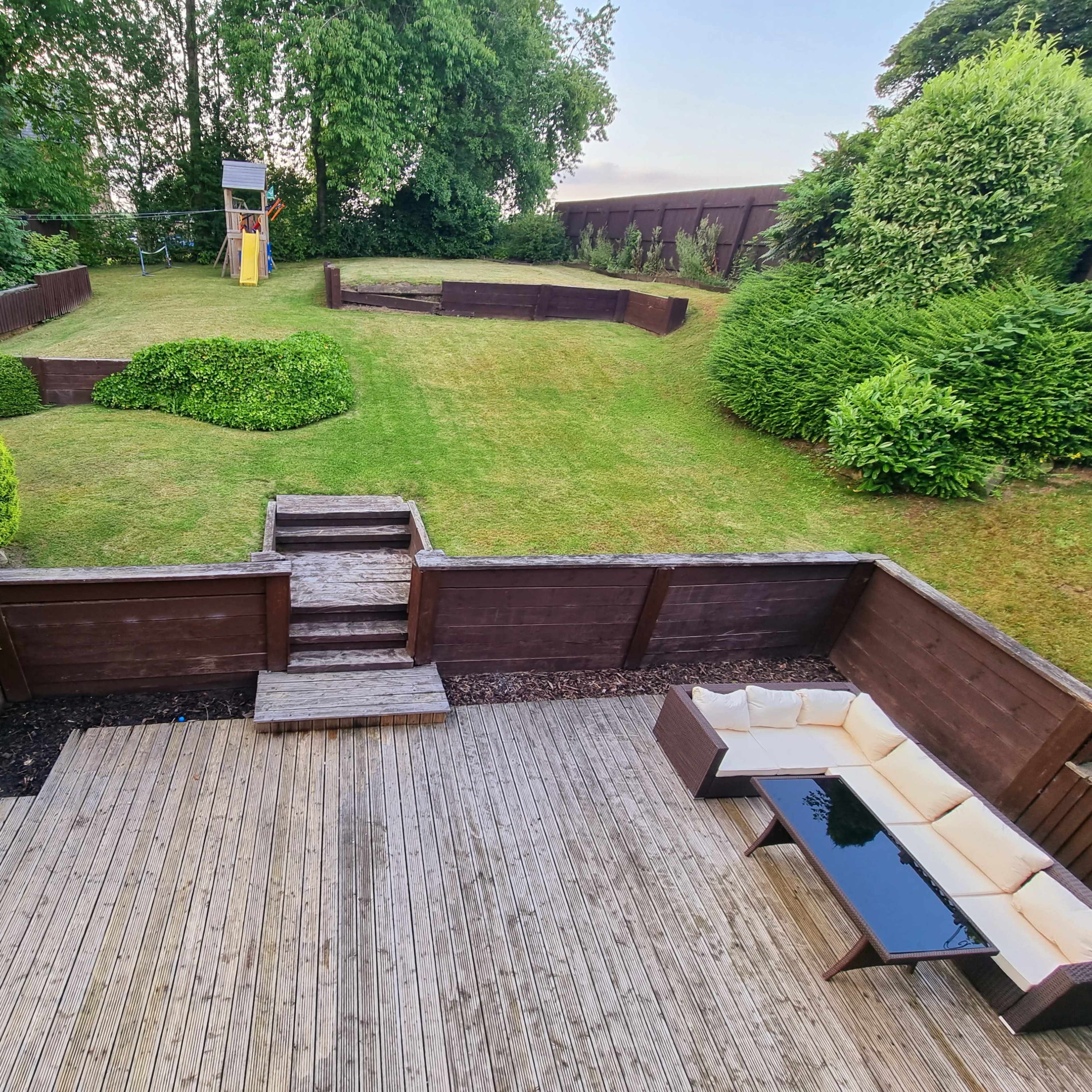 A landscaped garden with a grassy area, bordered by wooden walls and bushes, and a wooden deck with outdoor seating.