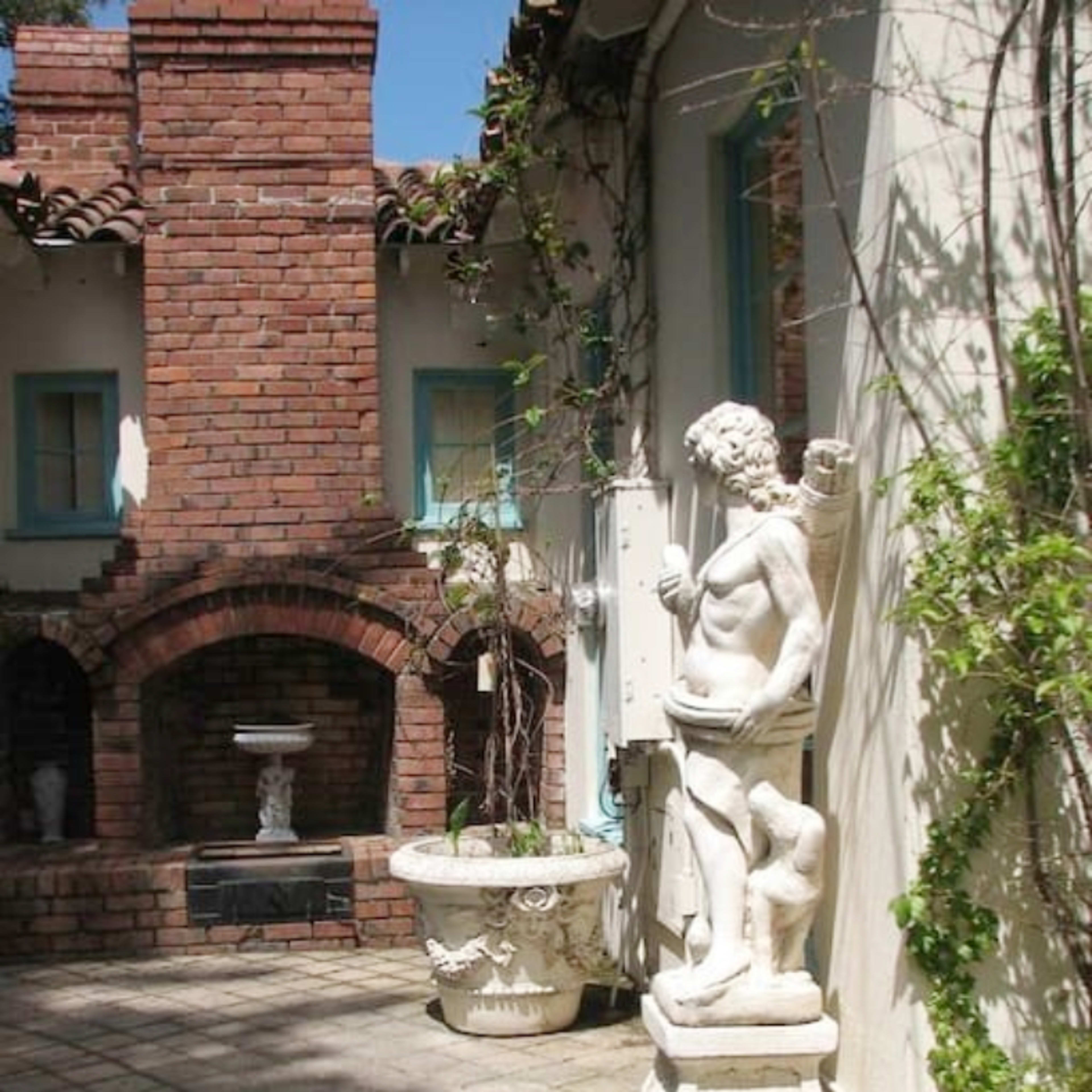A white statue of a muscular figure stands on a pedestal beside a potted plant in a brick courtyard.
