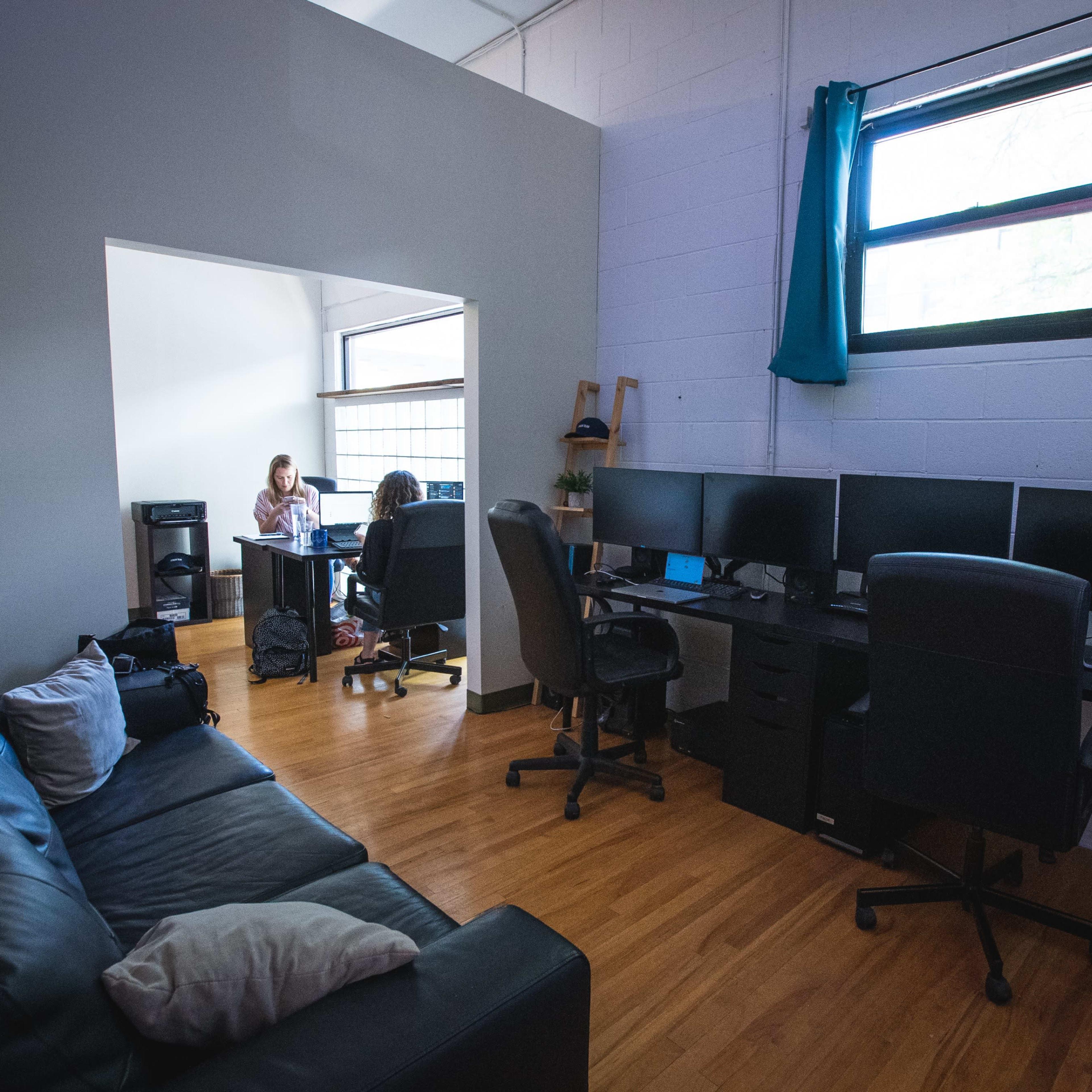 A shared office space with a couch on one side and multiple computer monitors arranged on a desk, while two women are working at a separate table in the background.