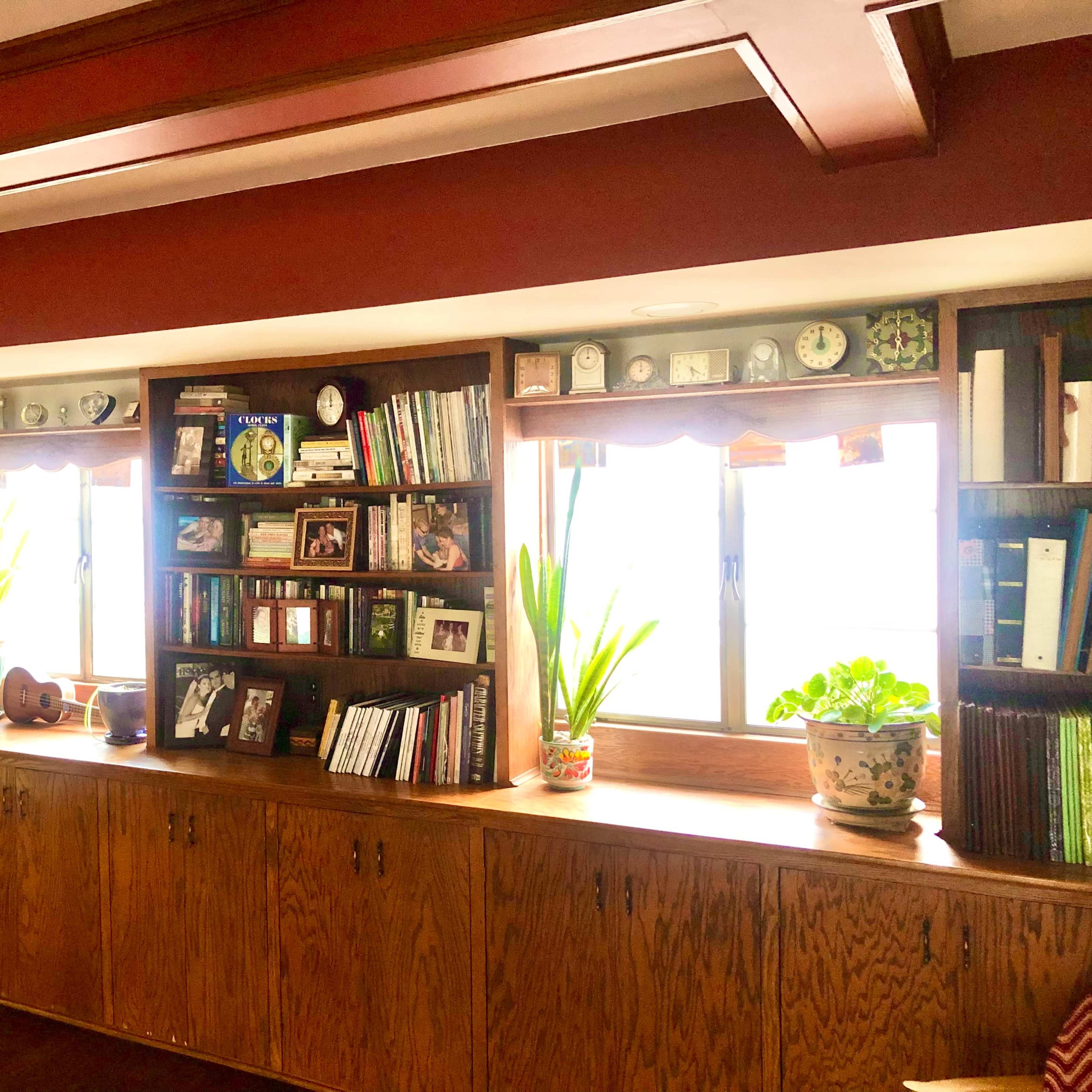 A wooden bookshelf with various books, framed photos, plants, and decorative items, positioned beneath two windows.