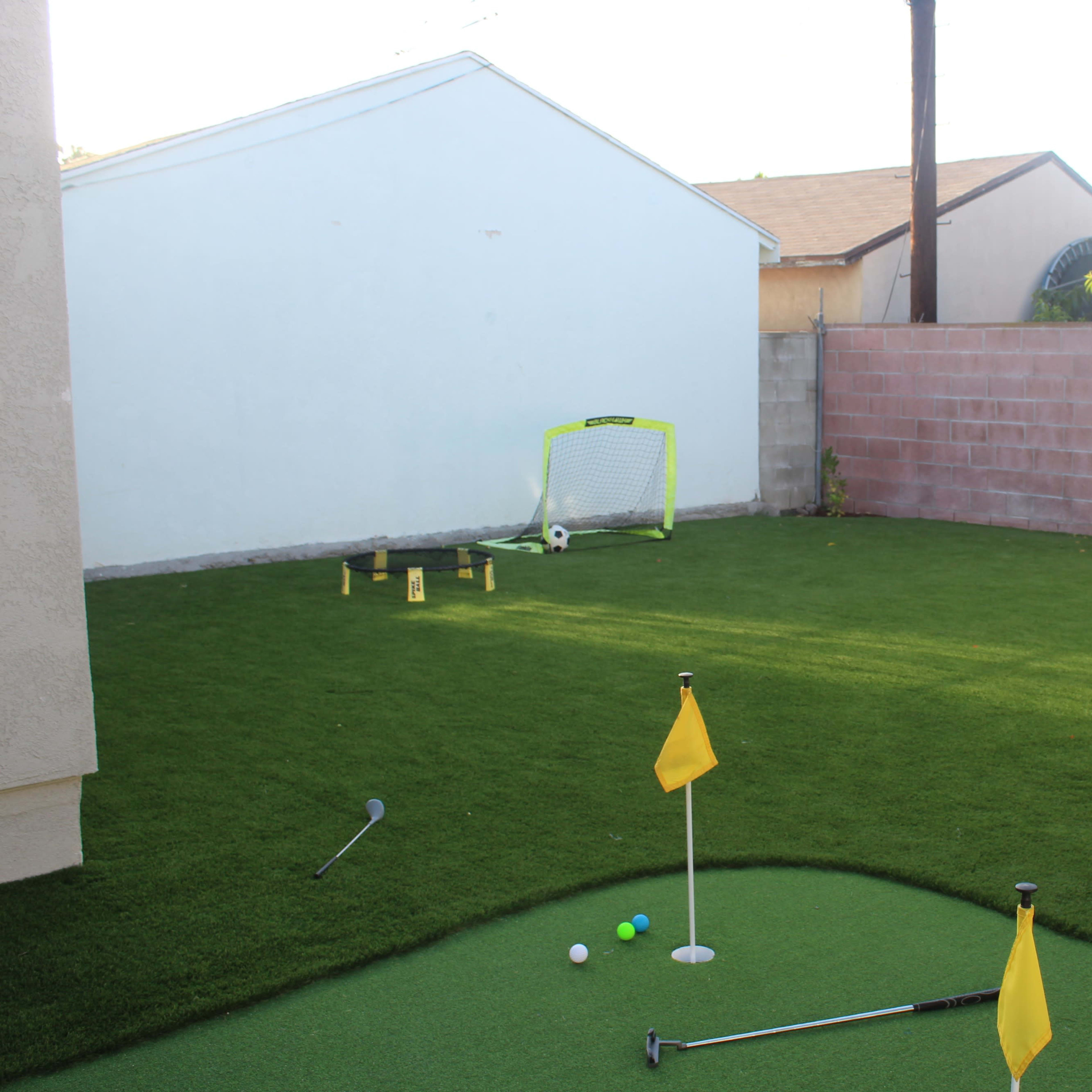 A small, landscaped backyard with artificial grass featuring a putting green, a soccer goal, and a few sports equipment scattered around.