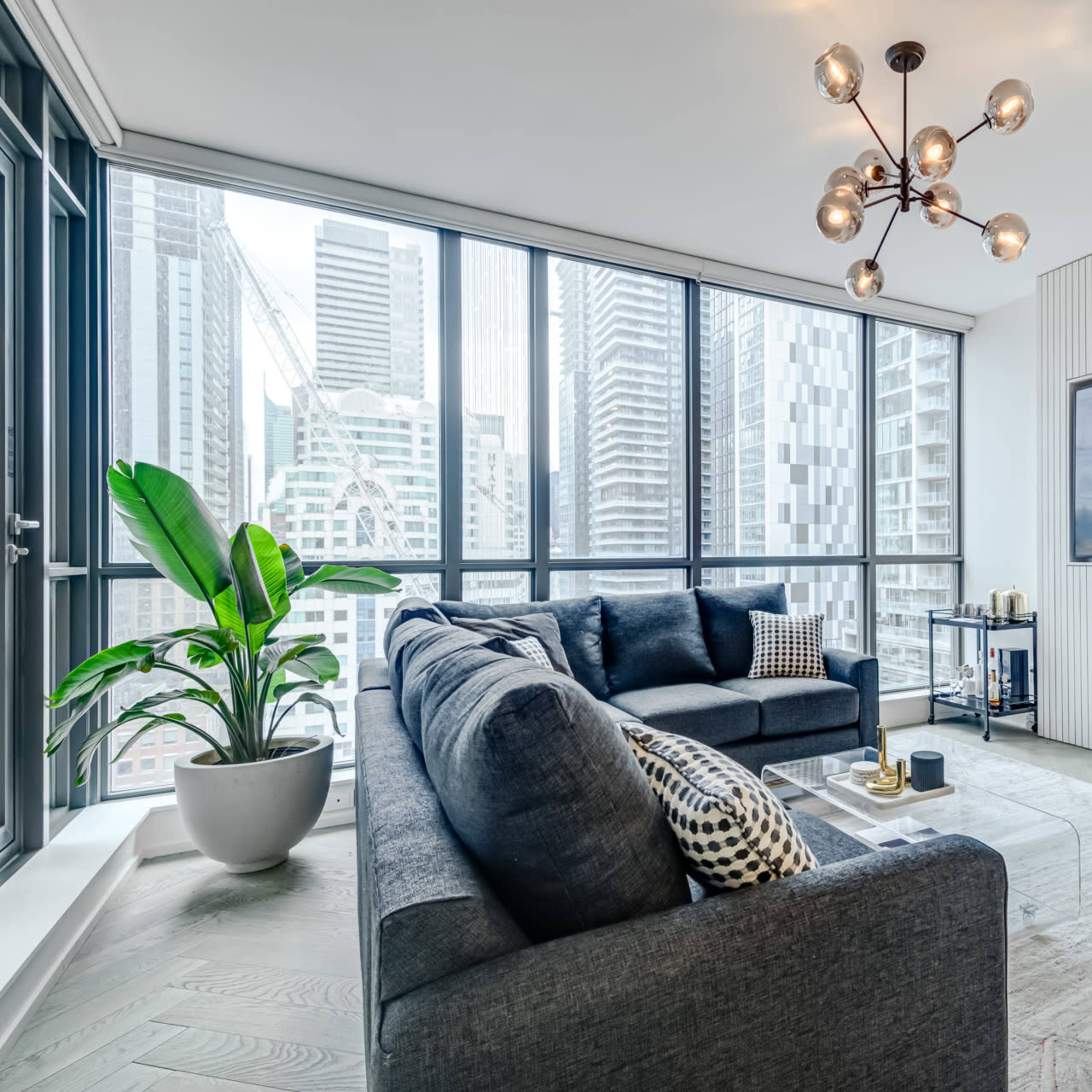A modern living room furnished with a gray sofa, a round chair, a large TV mounted on the wall, and a prominent window showcasing a cityscape.