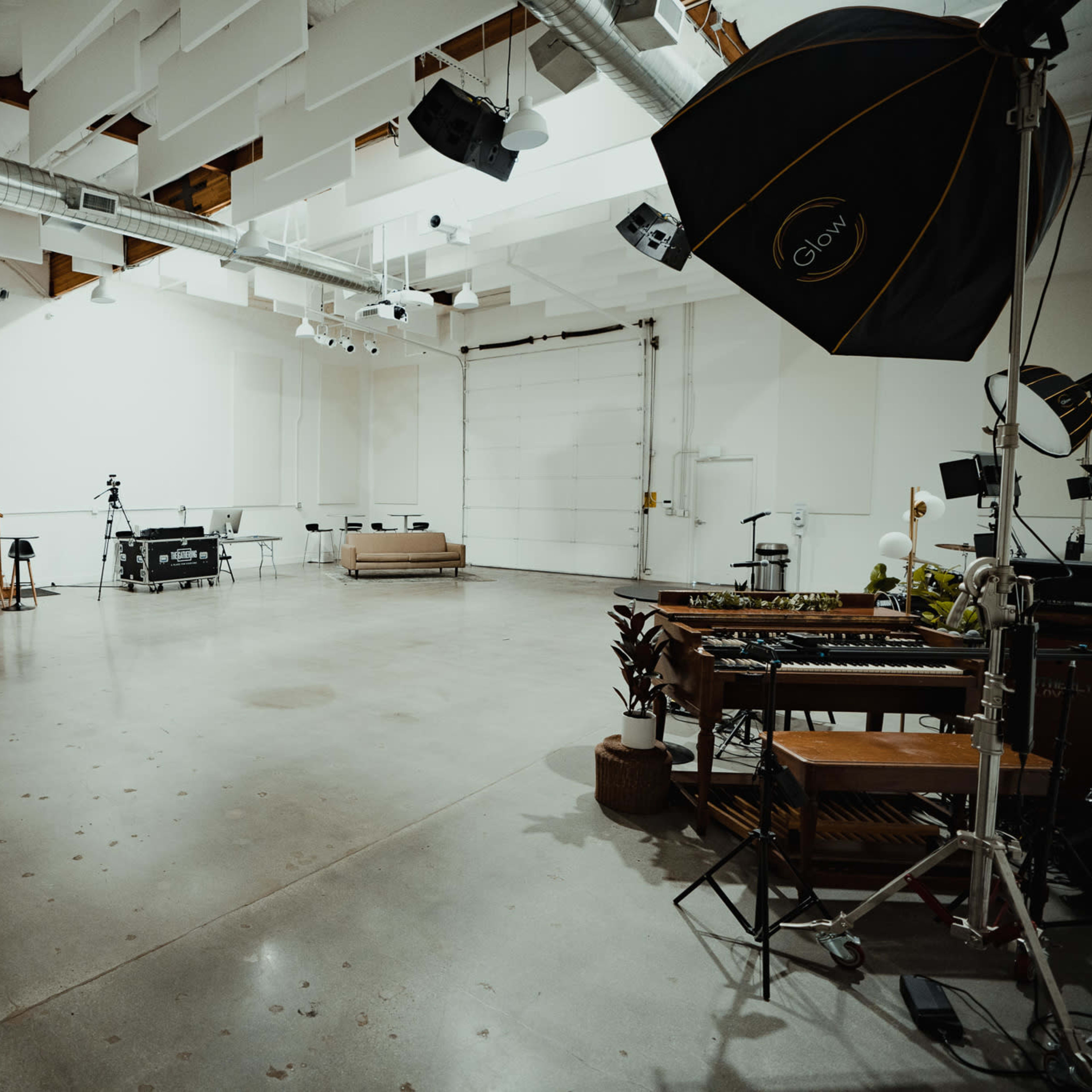 A spacious, well-lit studio features concrete floors, a staircase, and several photography and video equipment setups.