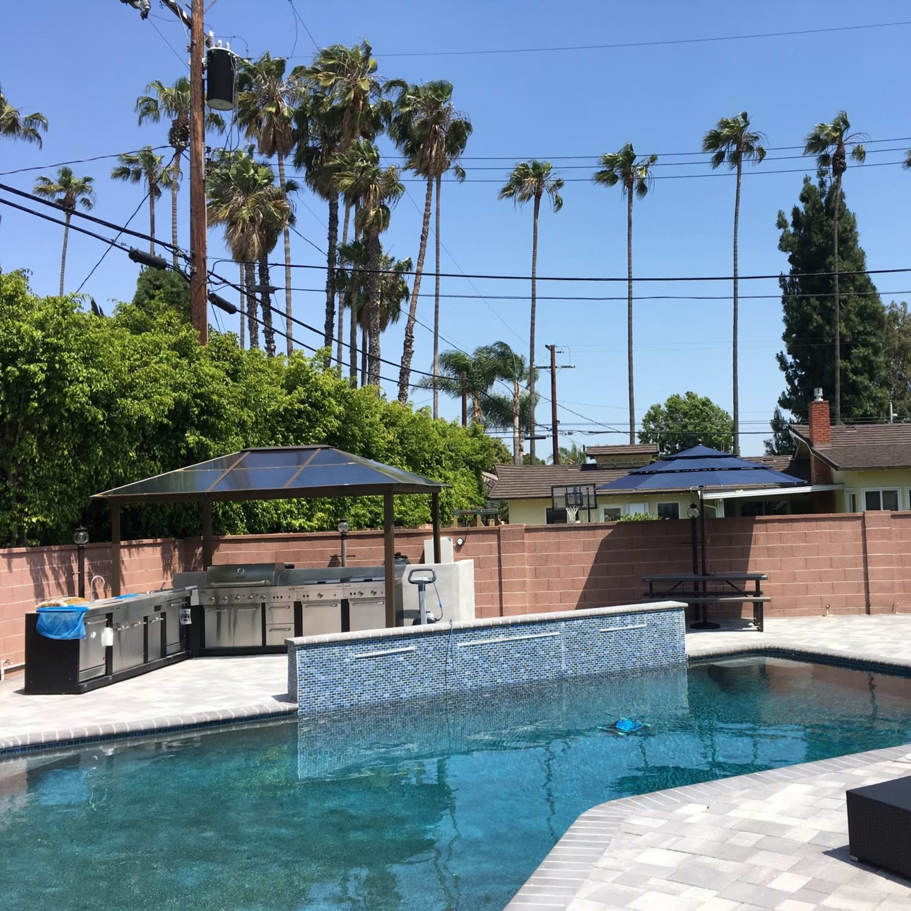 A backyard pool area features a patio with an outdoor kitchen, picnic tables, and palm trees in the background.
