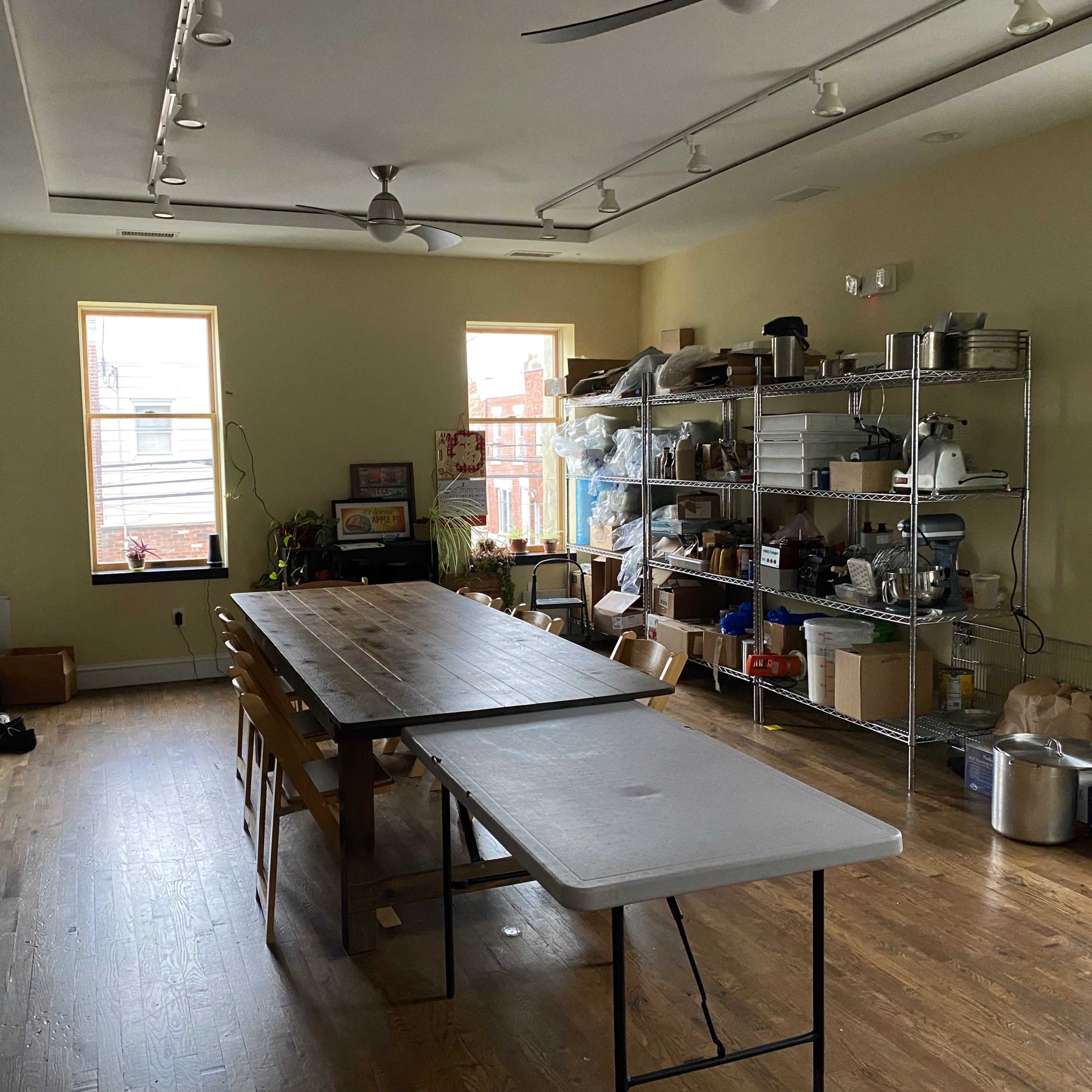 A spacious room with a long table in the center, surrounded by shelves filled with various kitchen items and plants near large windows.