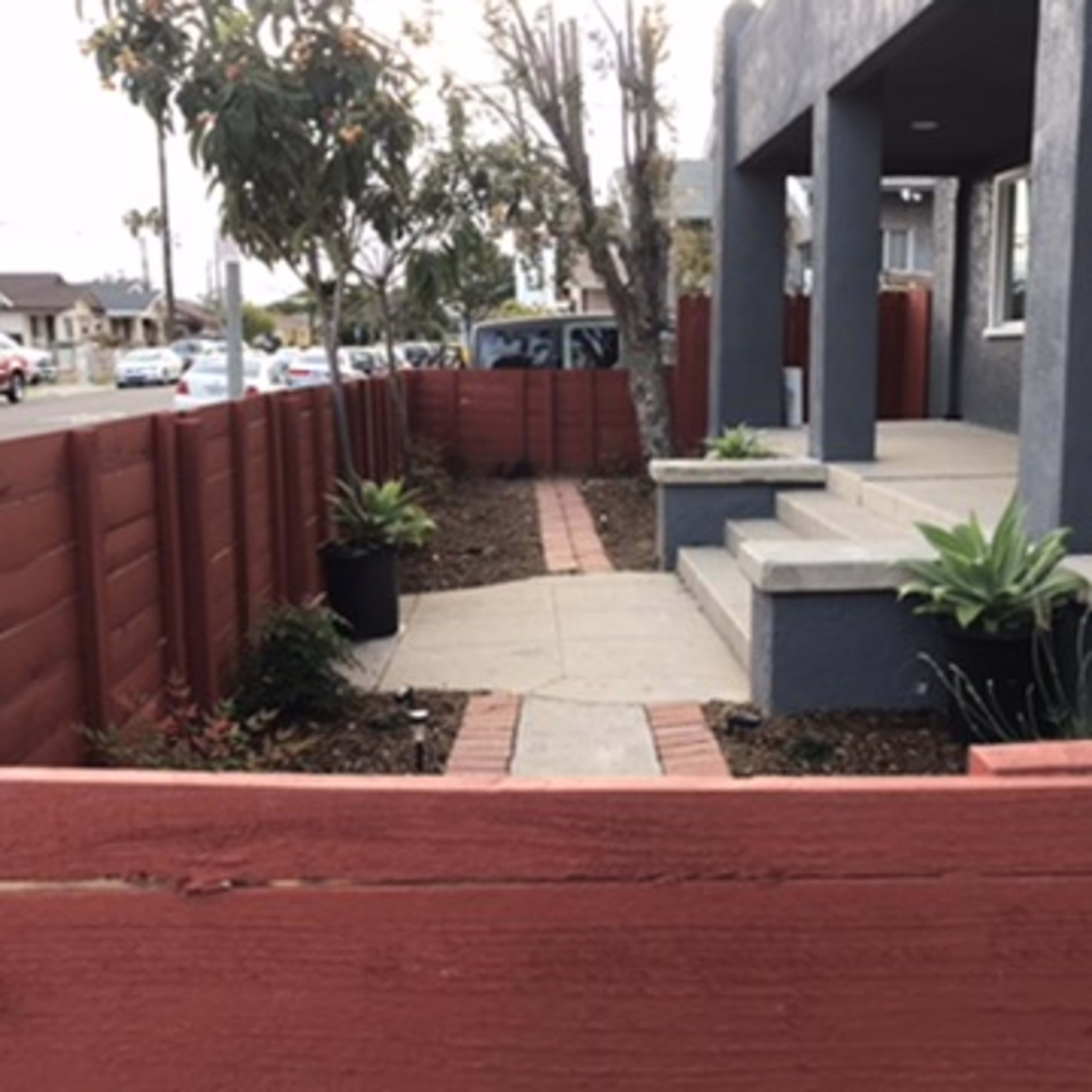 A walkway lined with plants leading to a house with a gray exterior and a red wooden fence.