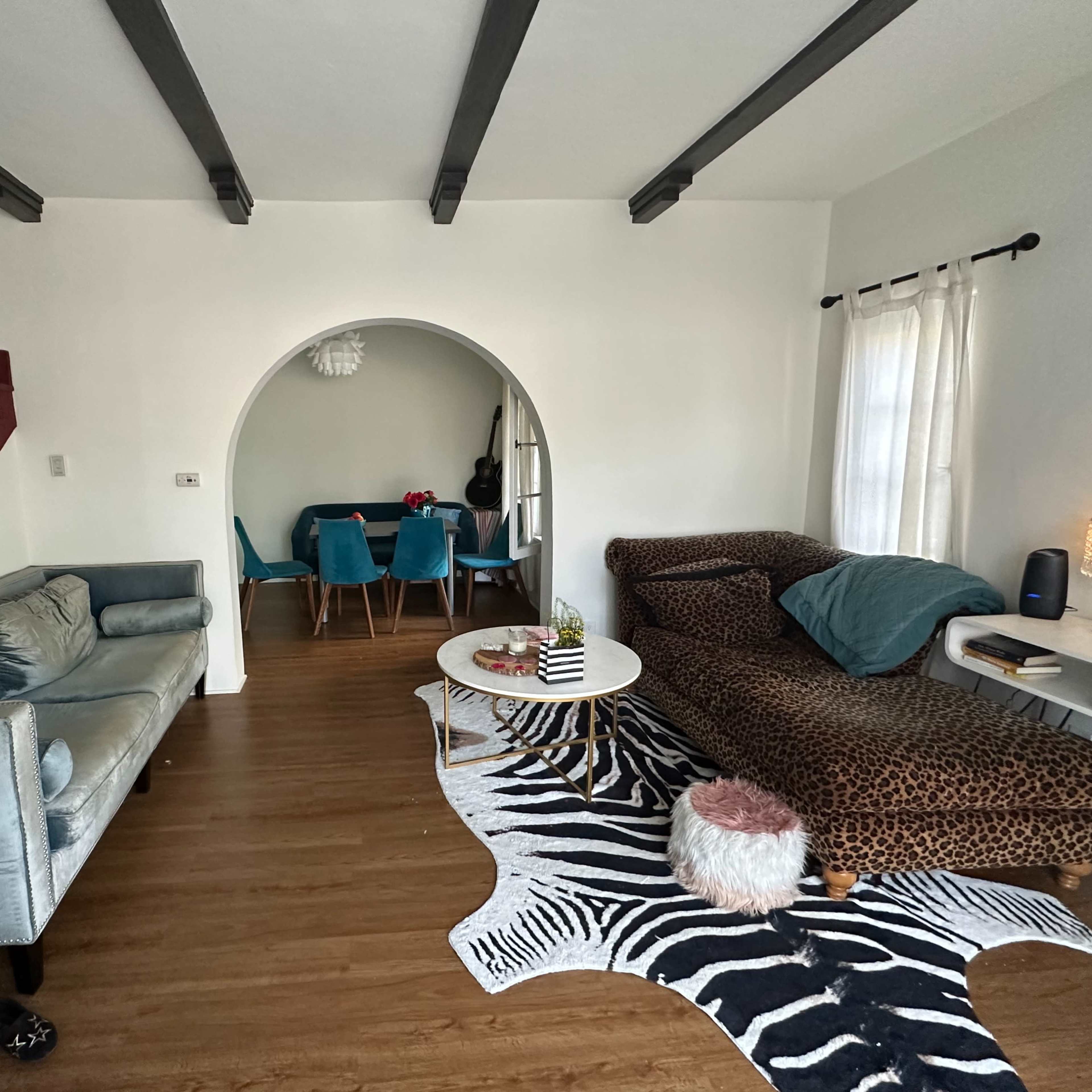 A living room featuring a blue velvet sofa, a leopard-print couch, a zebra-patterned rug, and a dining area with teal chairs visible through an archway.
