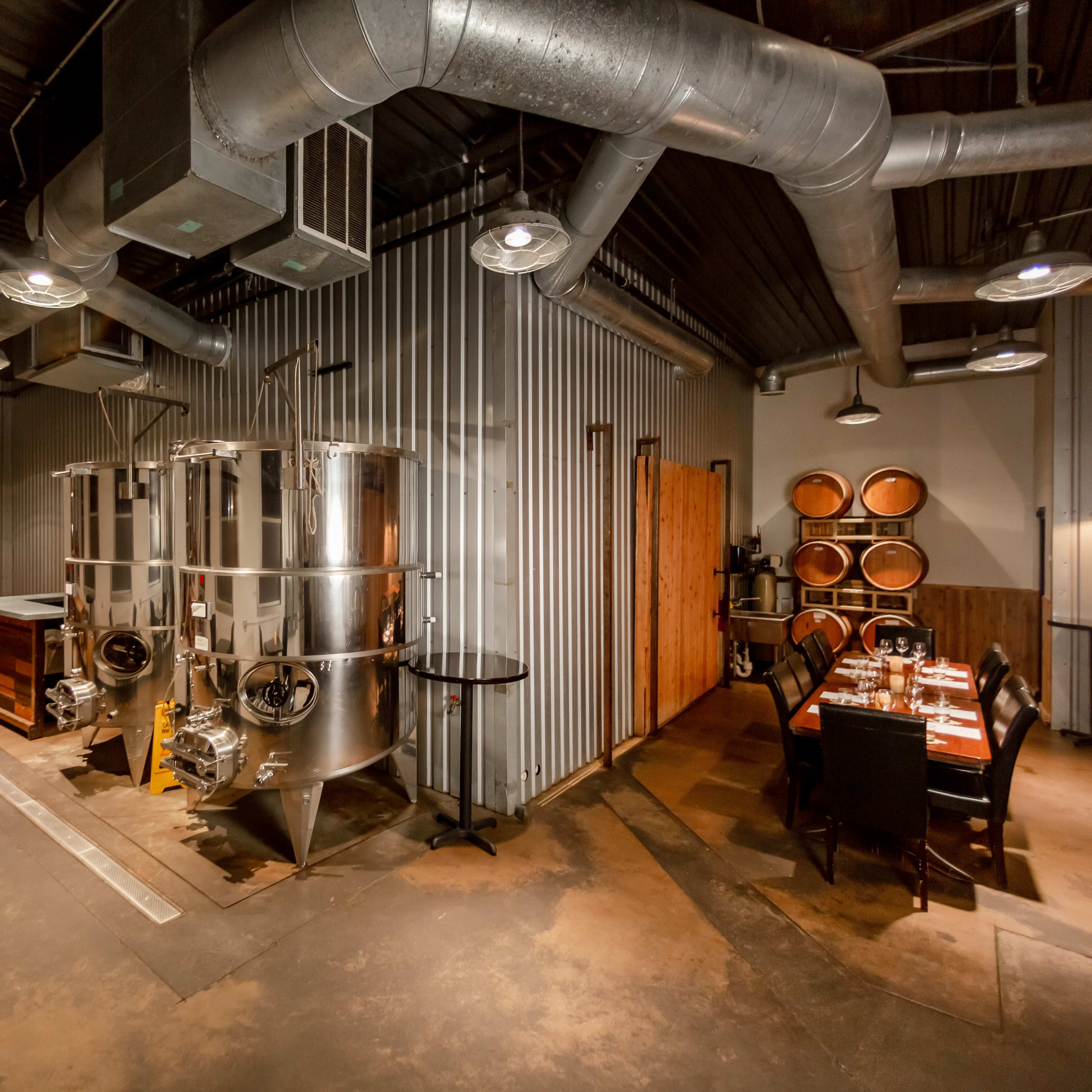 An industrial-style interior featuring metal fermentation tanks, wooden barrels, and a dining area with a table set for multiple guests.
