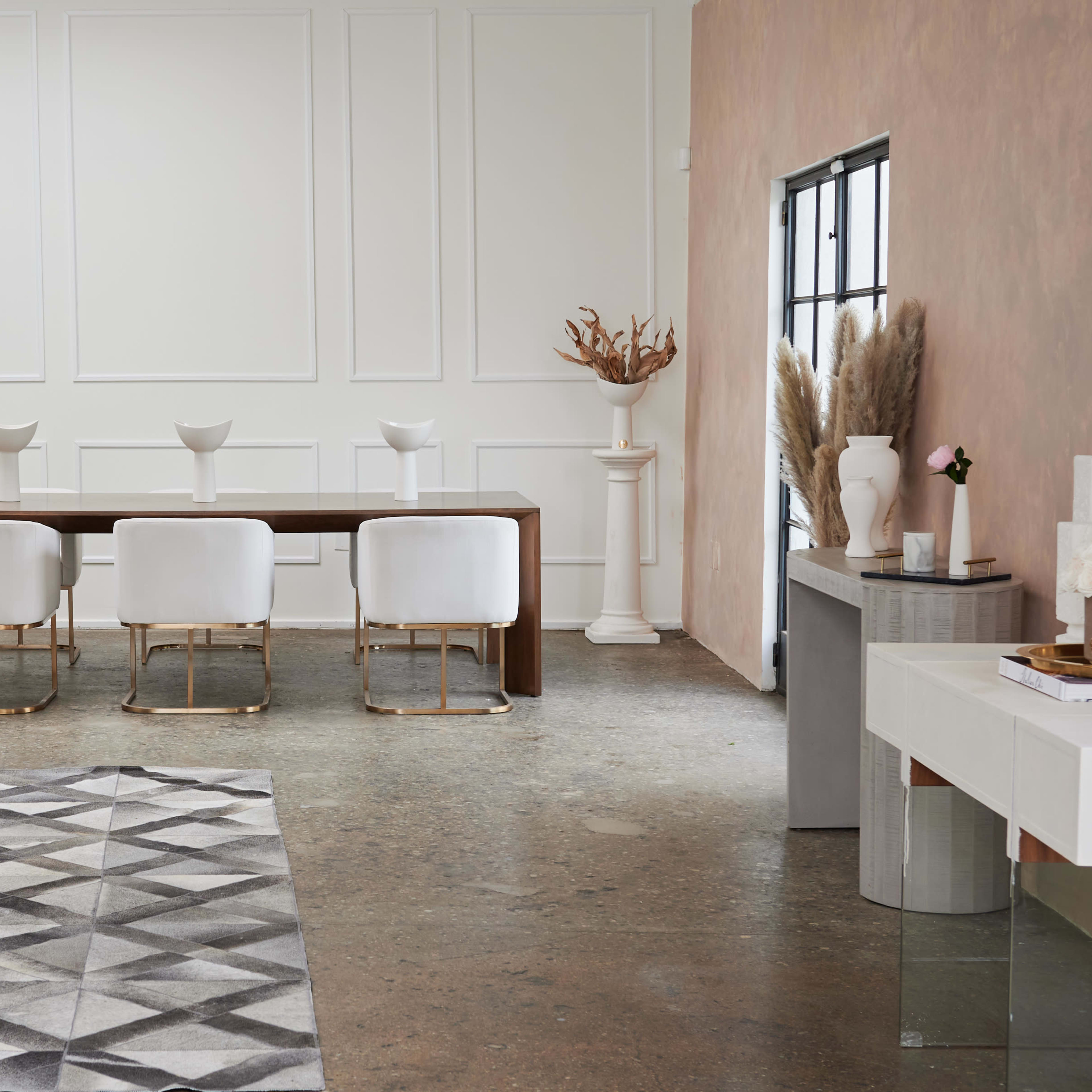 A modern dining area features a long wooden table surrounded by white chairs, with a geometric rug on the floor and minimalist decor along the walls.