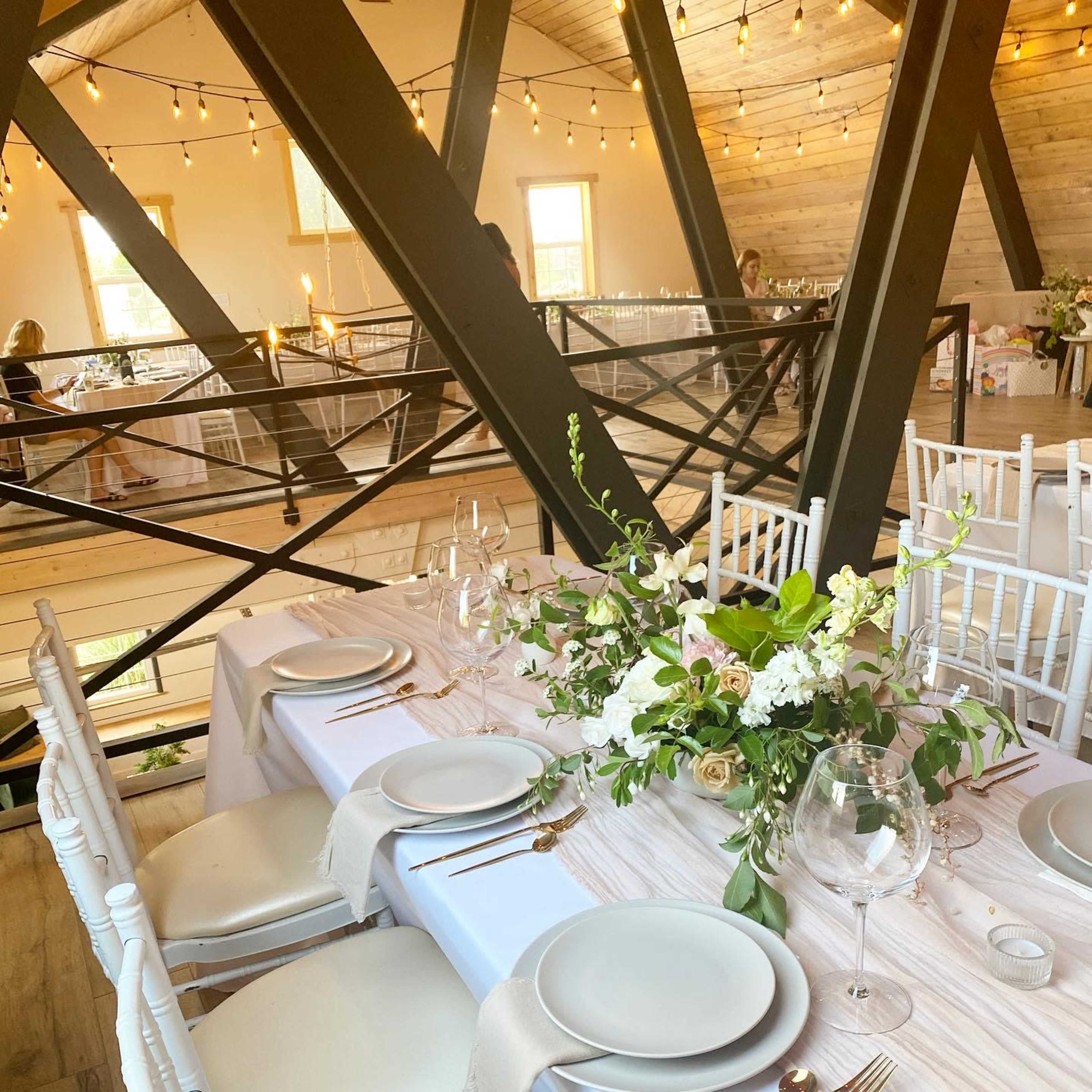 A beautifully arranged dining table with white chairs, adorned with floral centerpieces, set in an elegantly lit venue featuring wooden beams and string lights.
