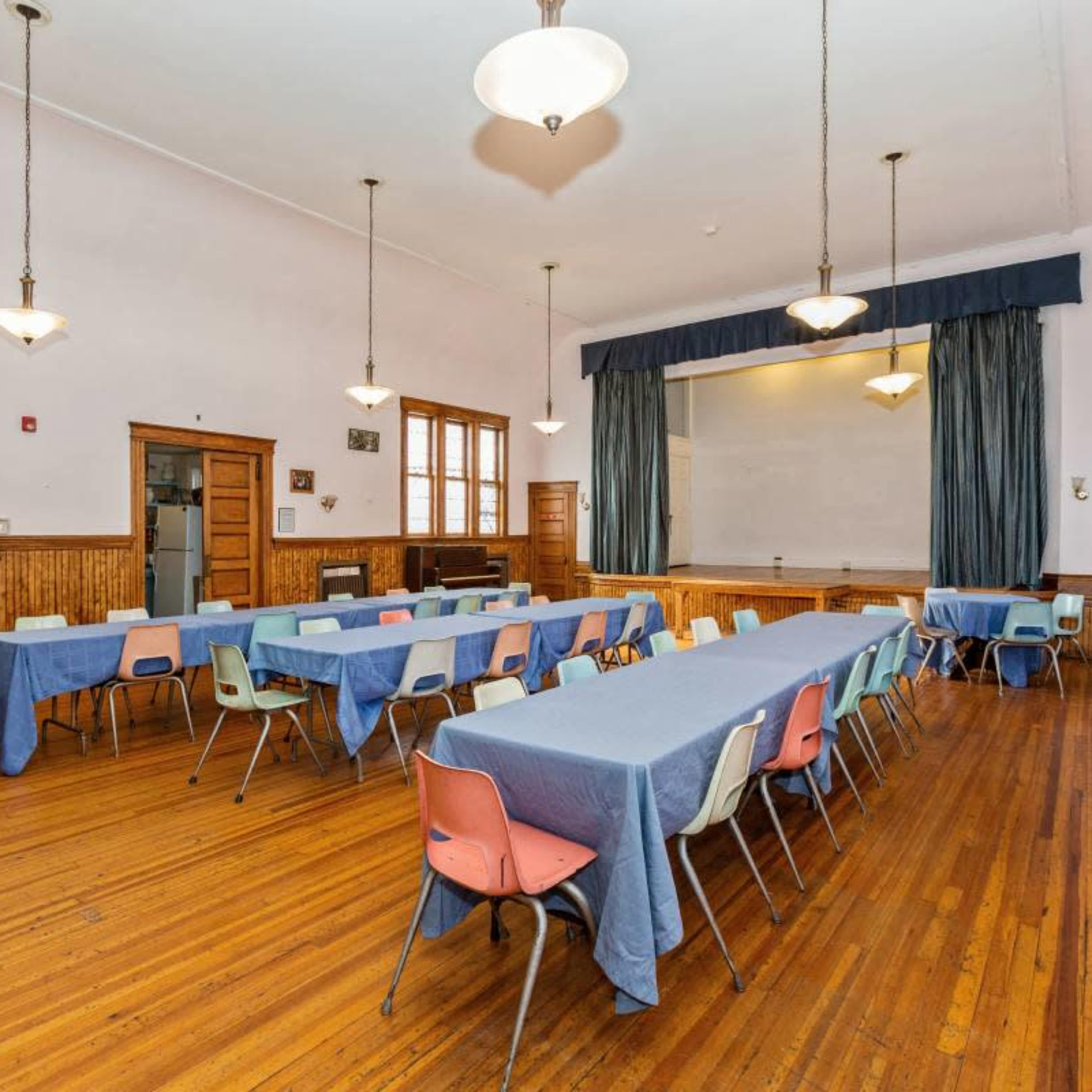 A spacious classroom or meeting room with several long tables covered in blue tablecloths and surrounded by chairs, set against a backdrop of wooden walls and a chalkboard.