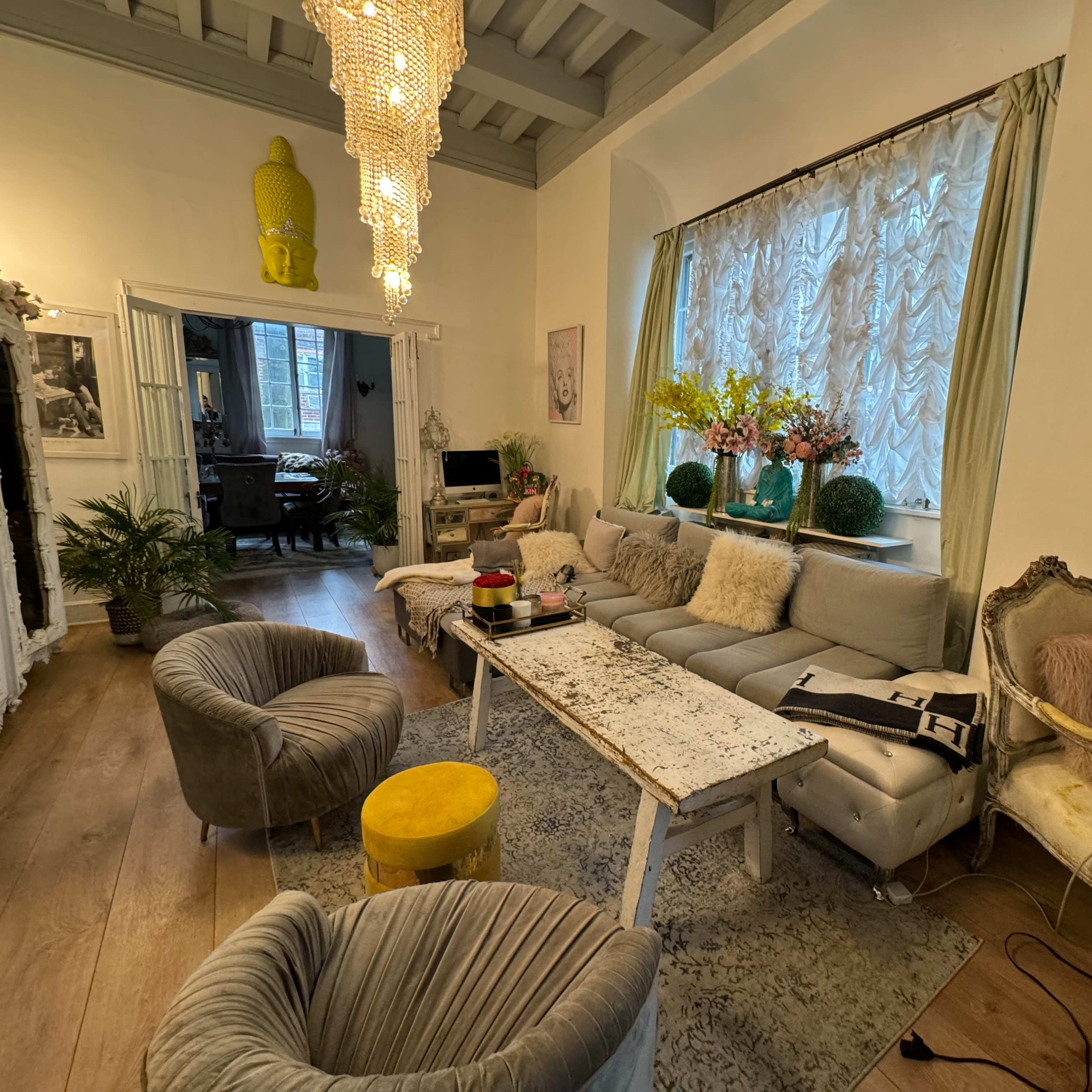 A cozy living room with a light-colored sofa, a distressed coffee table, and decorative plants, illuminated by a cluster of chandeliers.