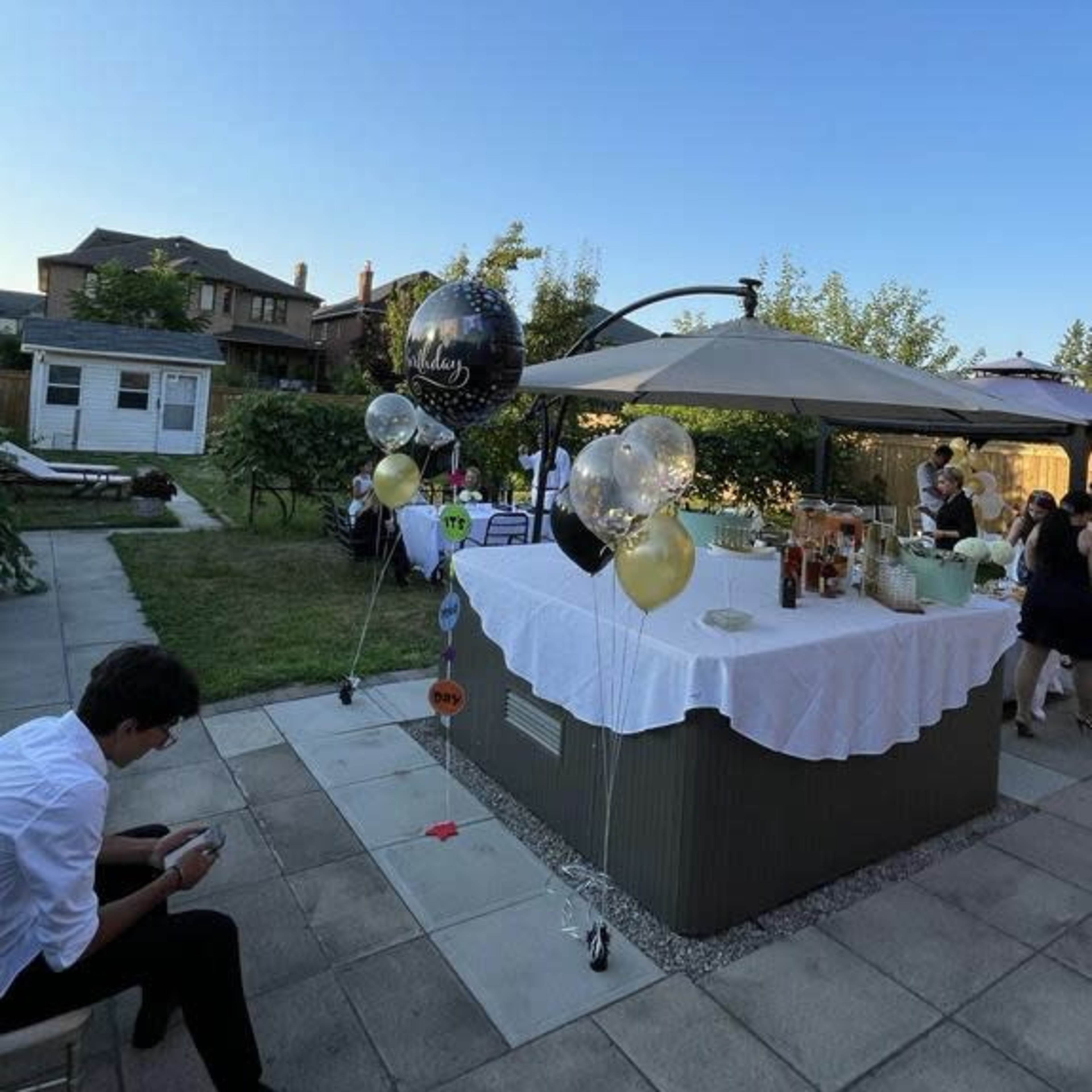 A garden setting features a table set with drinks and decorations, with guests socializing nearby under a clear blue sky.