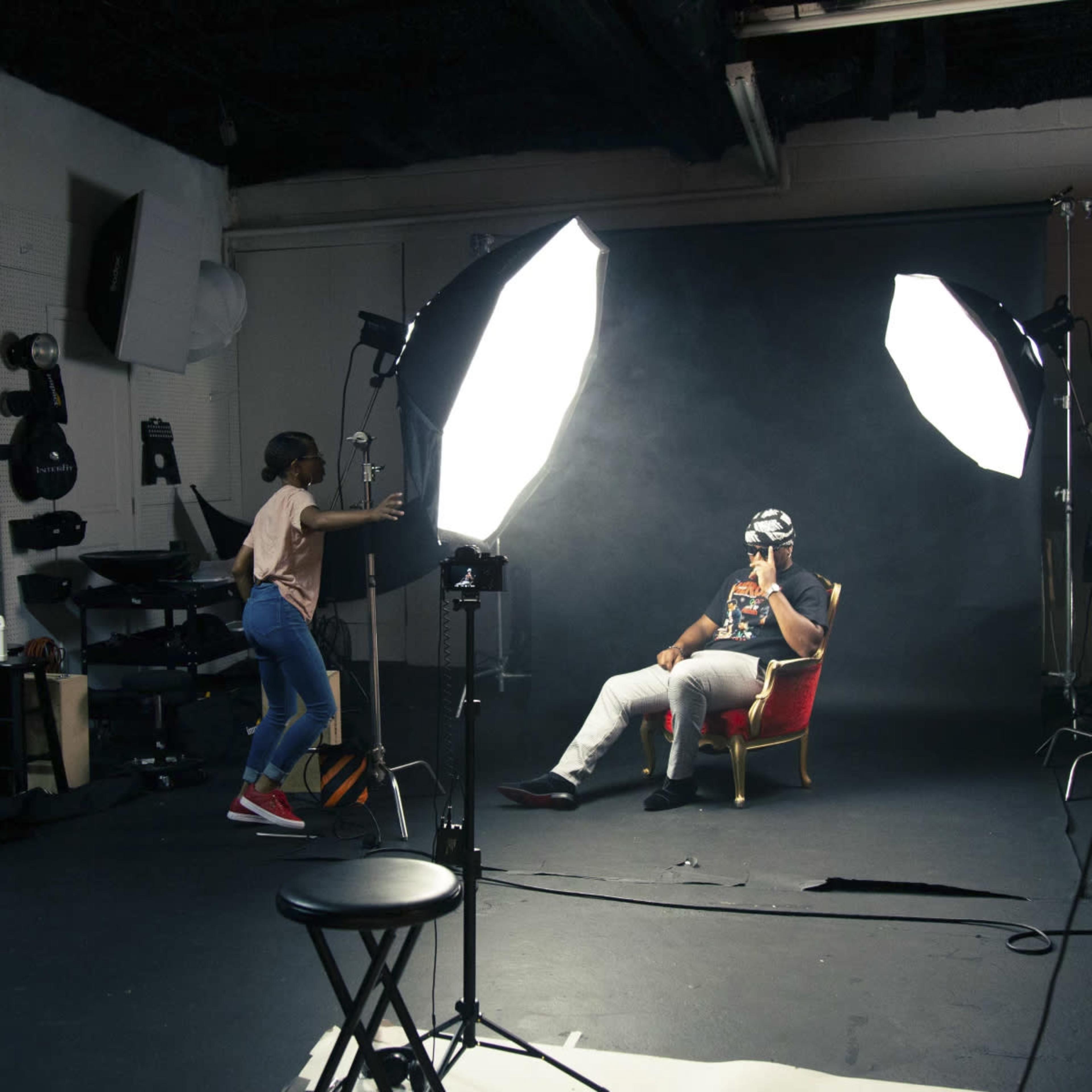 A person wearing a headset sits in a chair in a photo studio, while another individual adjusts lighting equipment nearby.