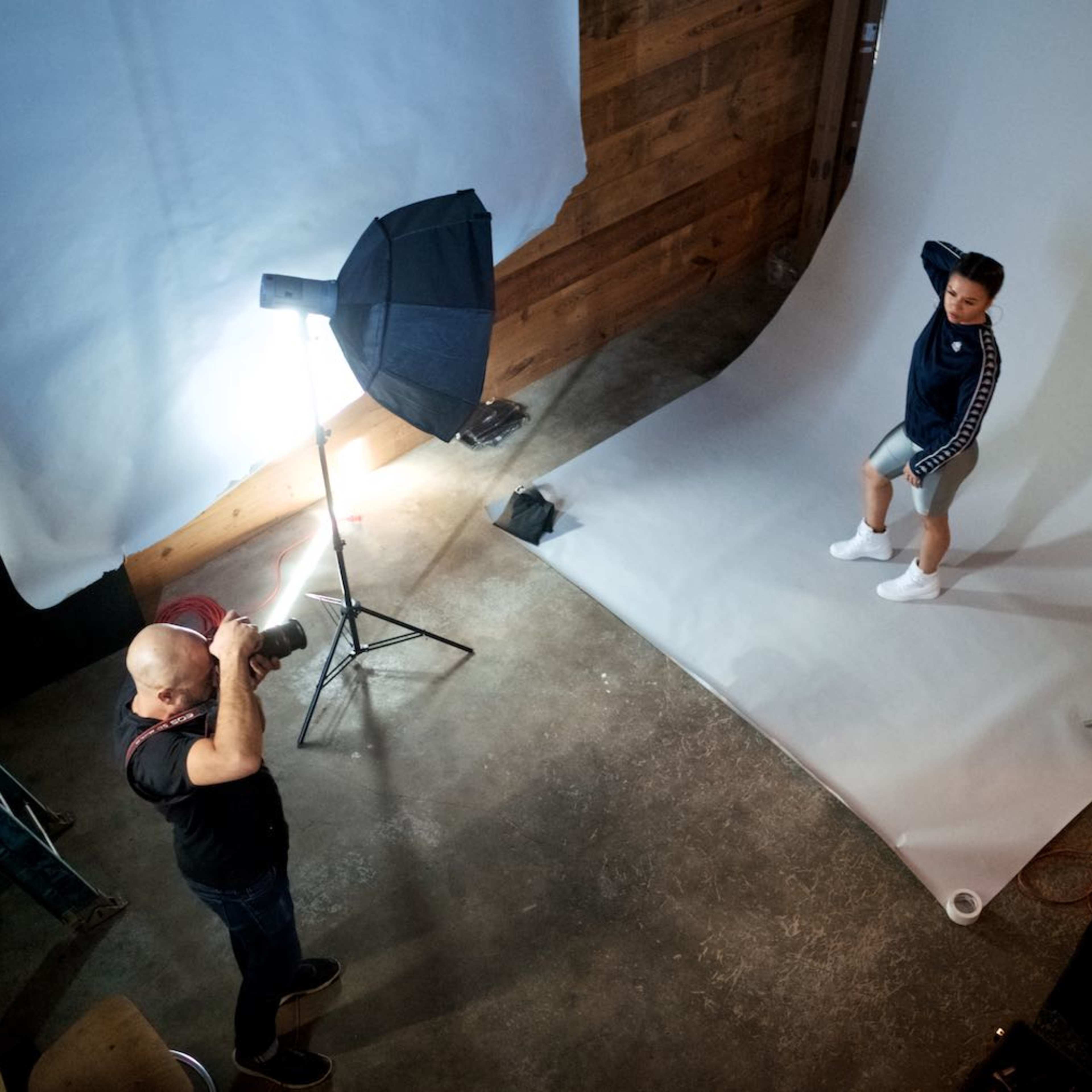 A photographer is capturing a model posing on a gray backdrop in a studio setting.