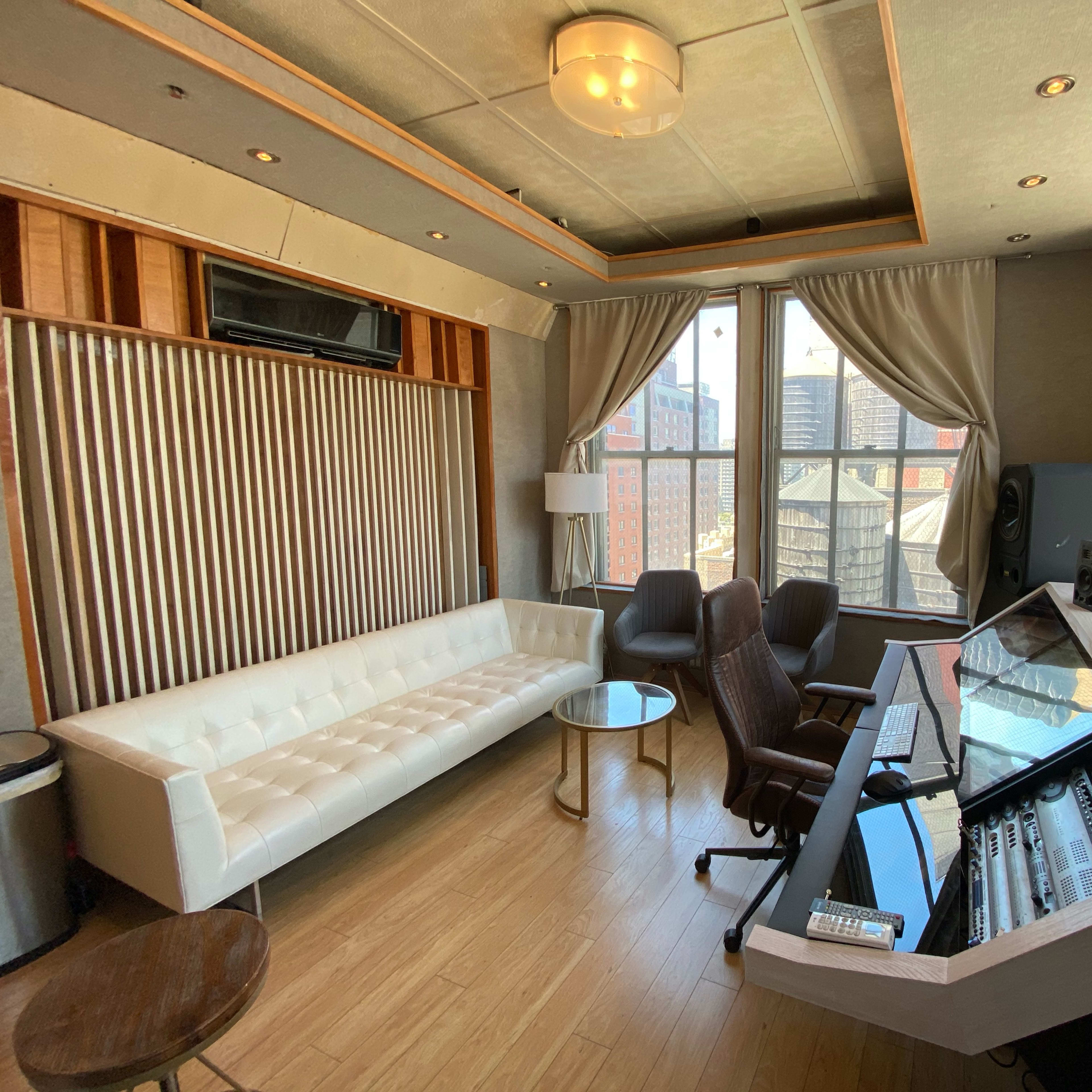 A modern recording studio with a mixing console, a white sofa, and large windows overlooking a cityscape.