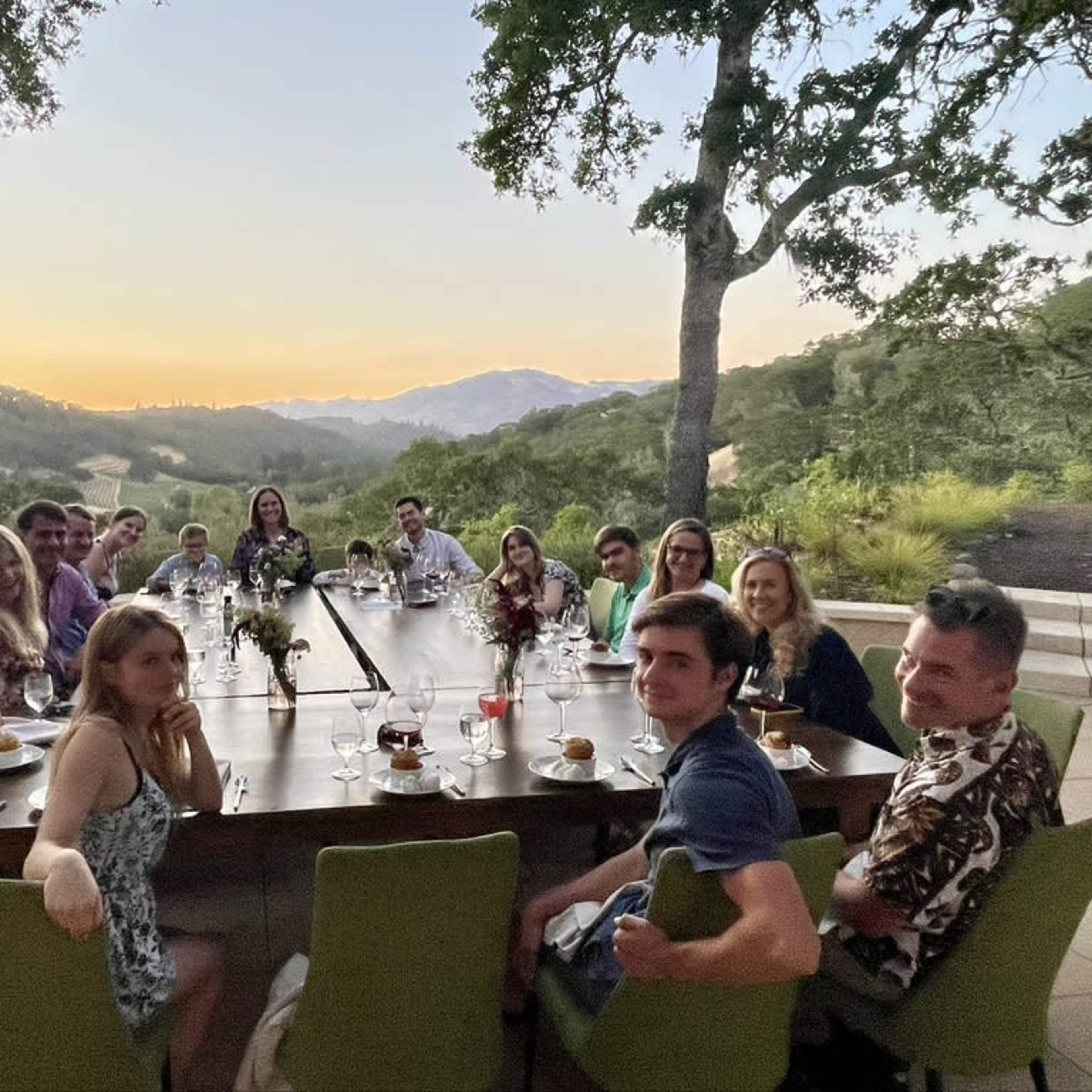 A group of people is seated around a long table outdoors, with a scenic view of hills and a sunset in the background.