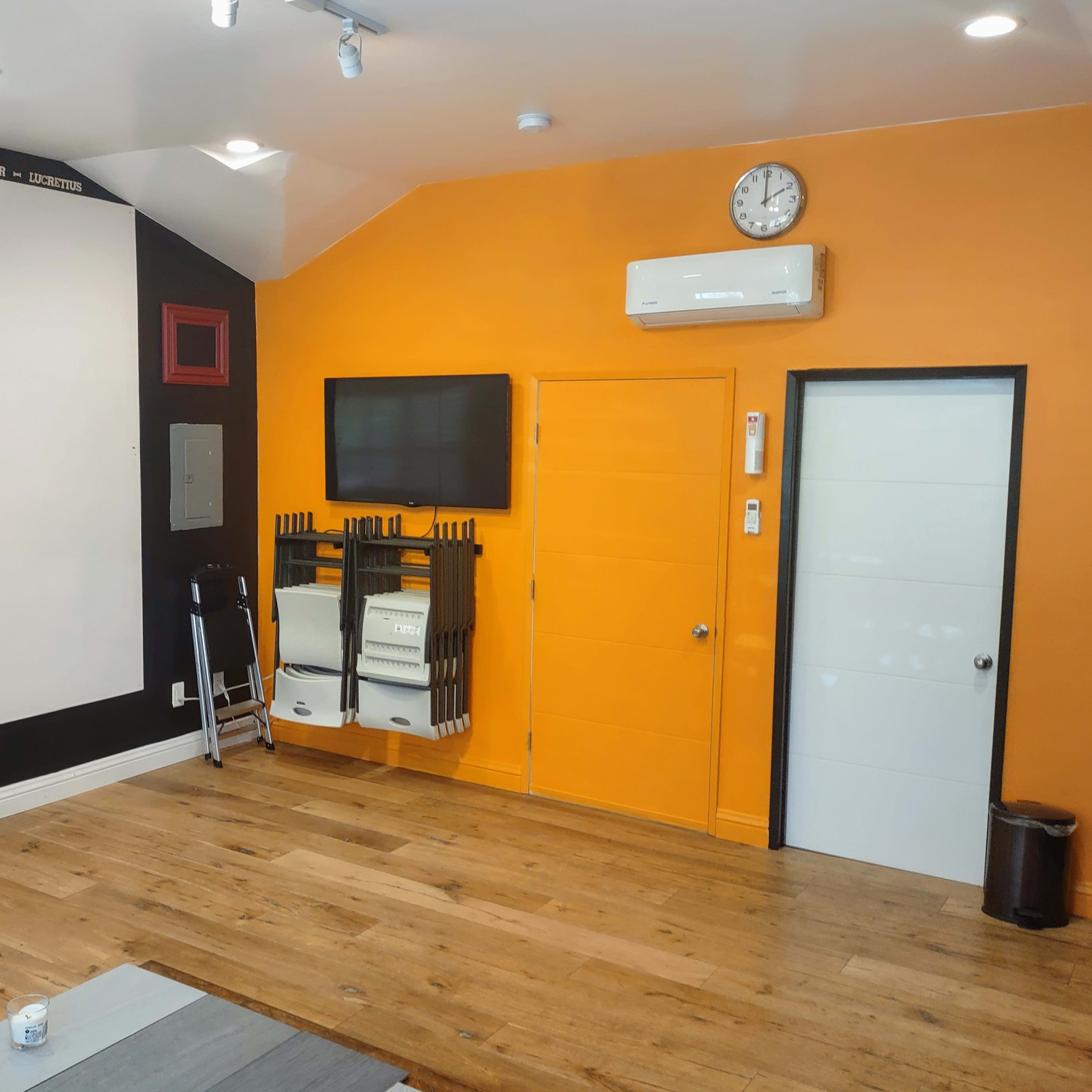 A brightly colored room with an orange accent wall, a whiteboard, wooden flooring, and a mounted TV along with a door leading to another space.