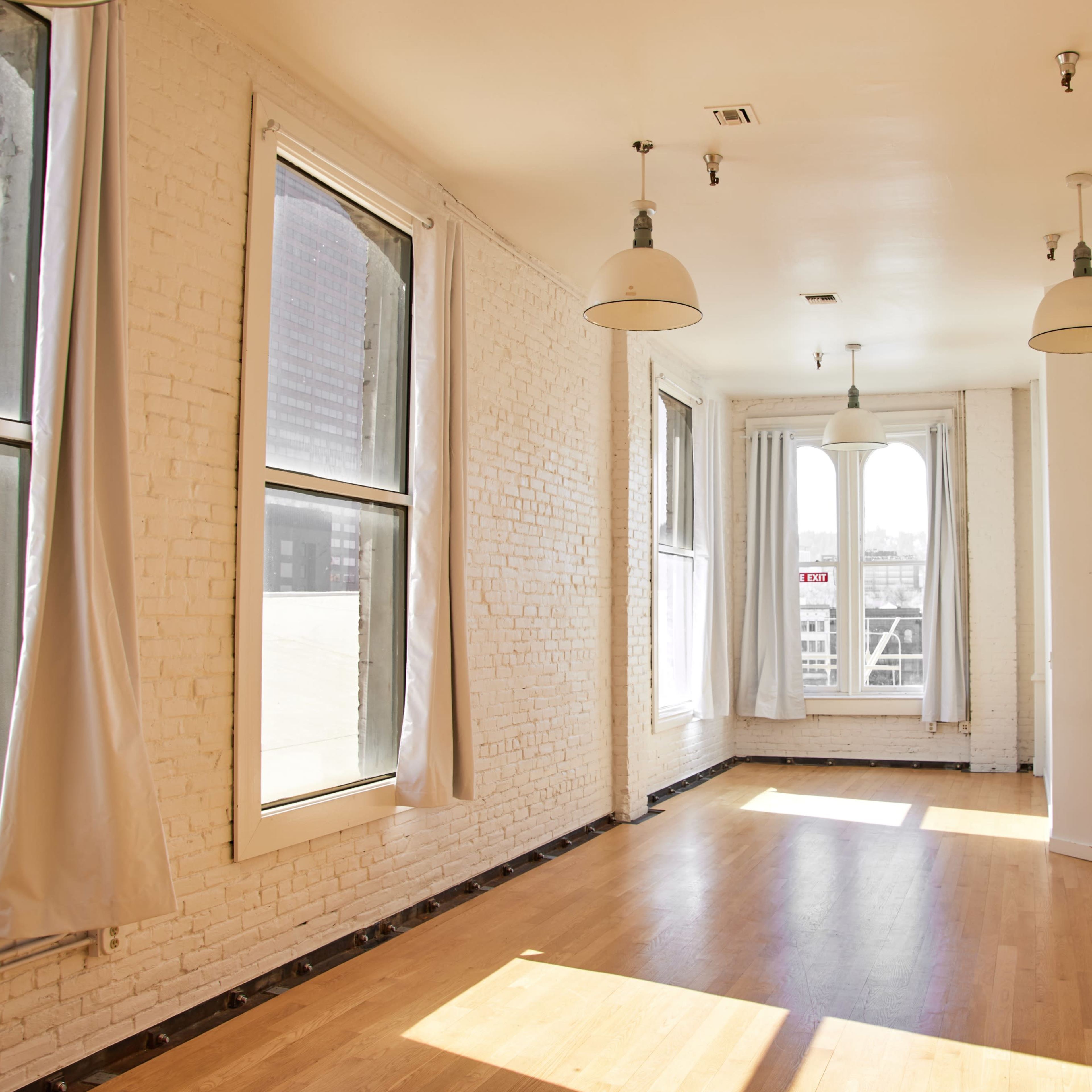 A spacious, sunlit room with large windows and wooden flooring, featuring exposed brick walls and industrial-style pendant lights.